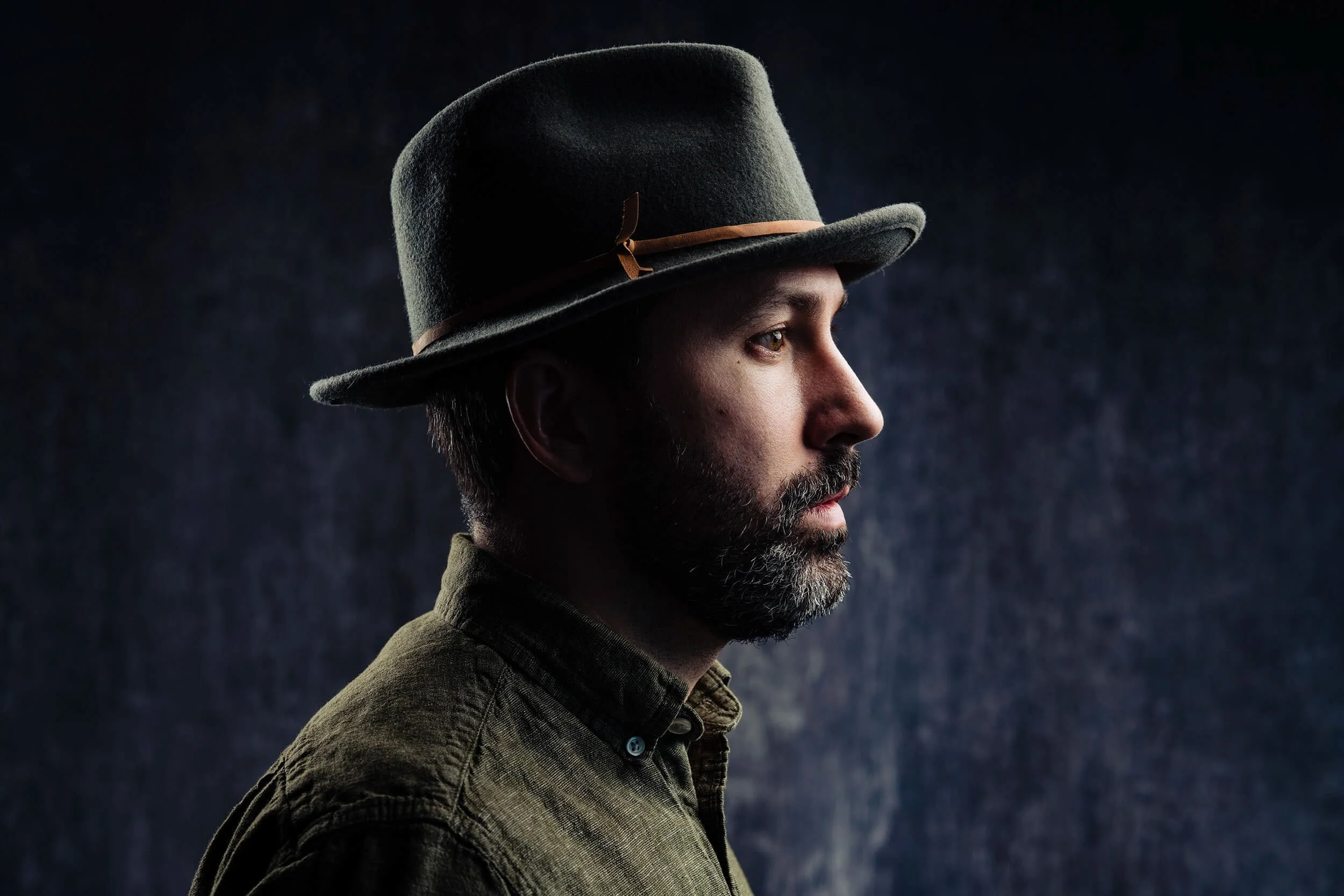 Side profile of a man wearing a greenish-brown button-up shirt and a gray felt fedora with a brown band, against a dark textured background.