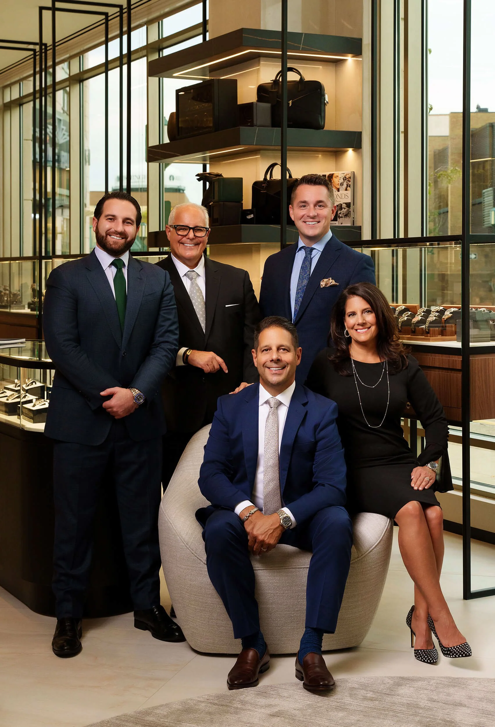 Group of six professionally dressed people posing inside a stylish watch or jewelry store with display cases and shelves filled with accessories.