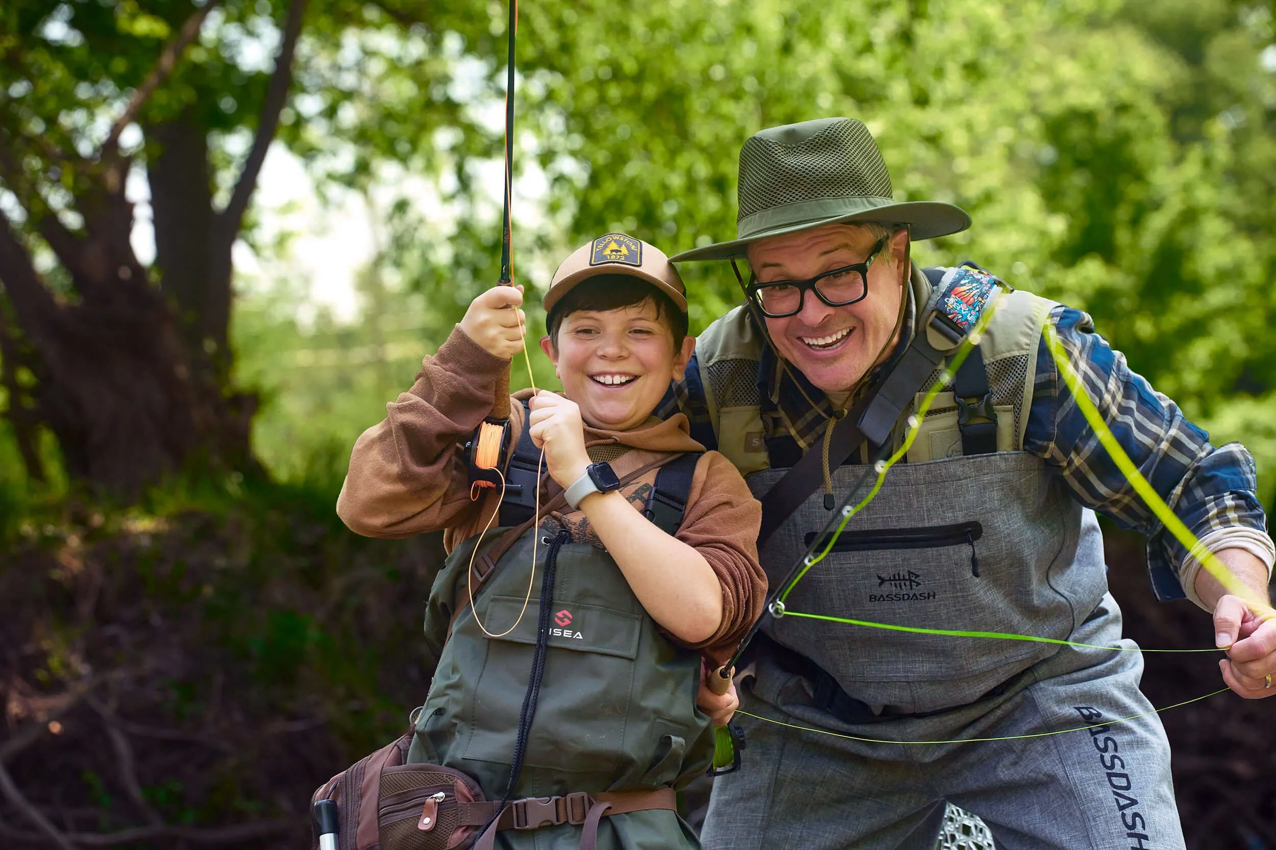 An older man and a young boy smiling while fishing in a forested area, with the boy holding a fishing rod.