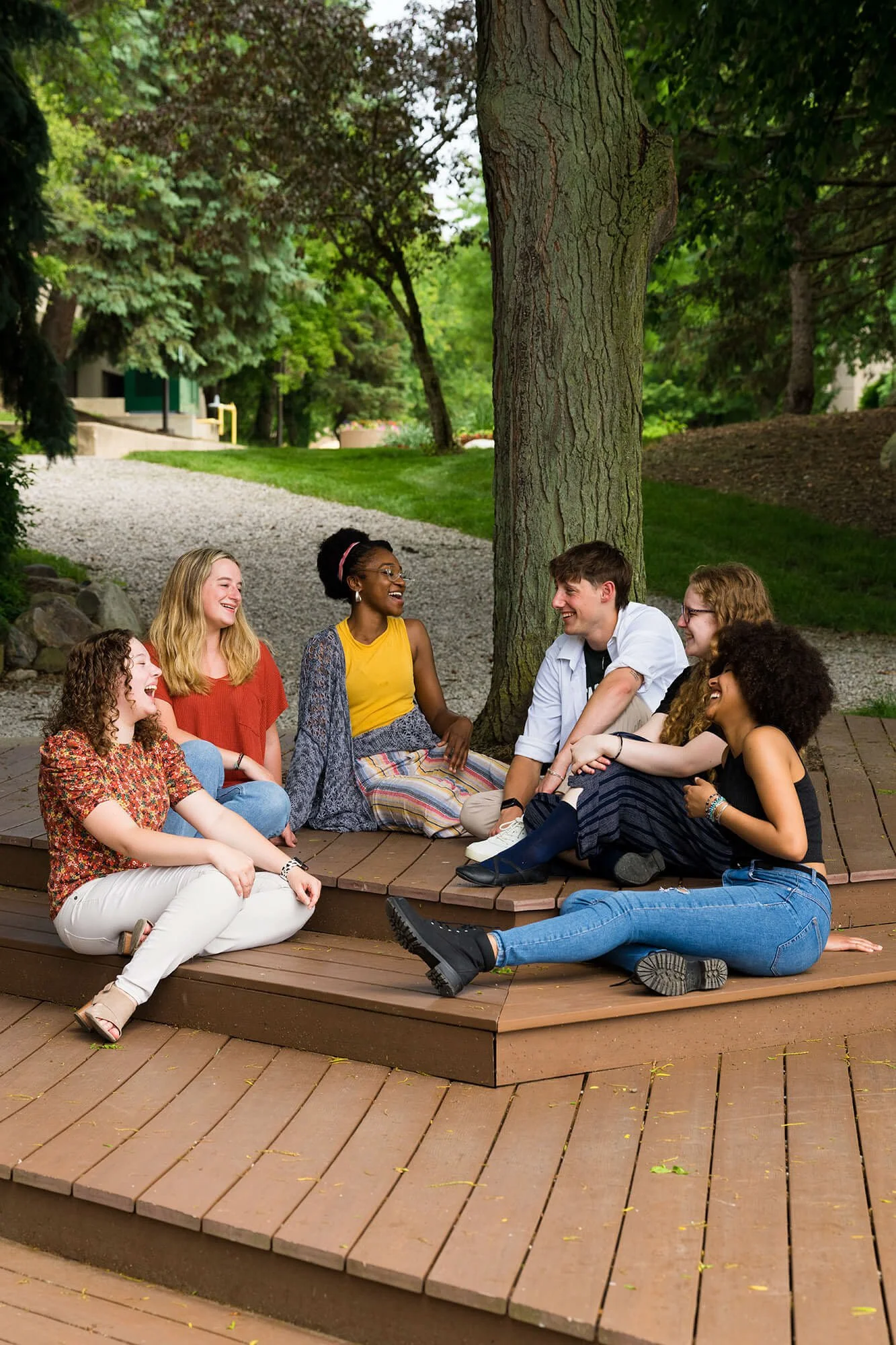 Group of seven diverse young people sitting and laughing together on a wooden circular platform outdoors in a park with green trees and grass.