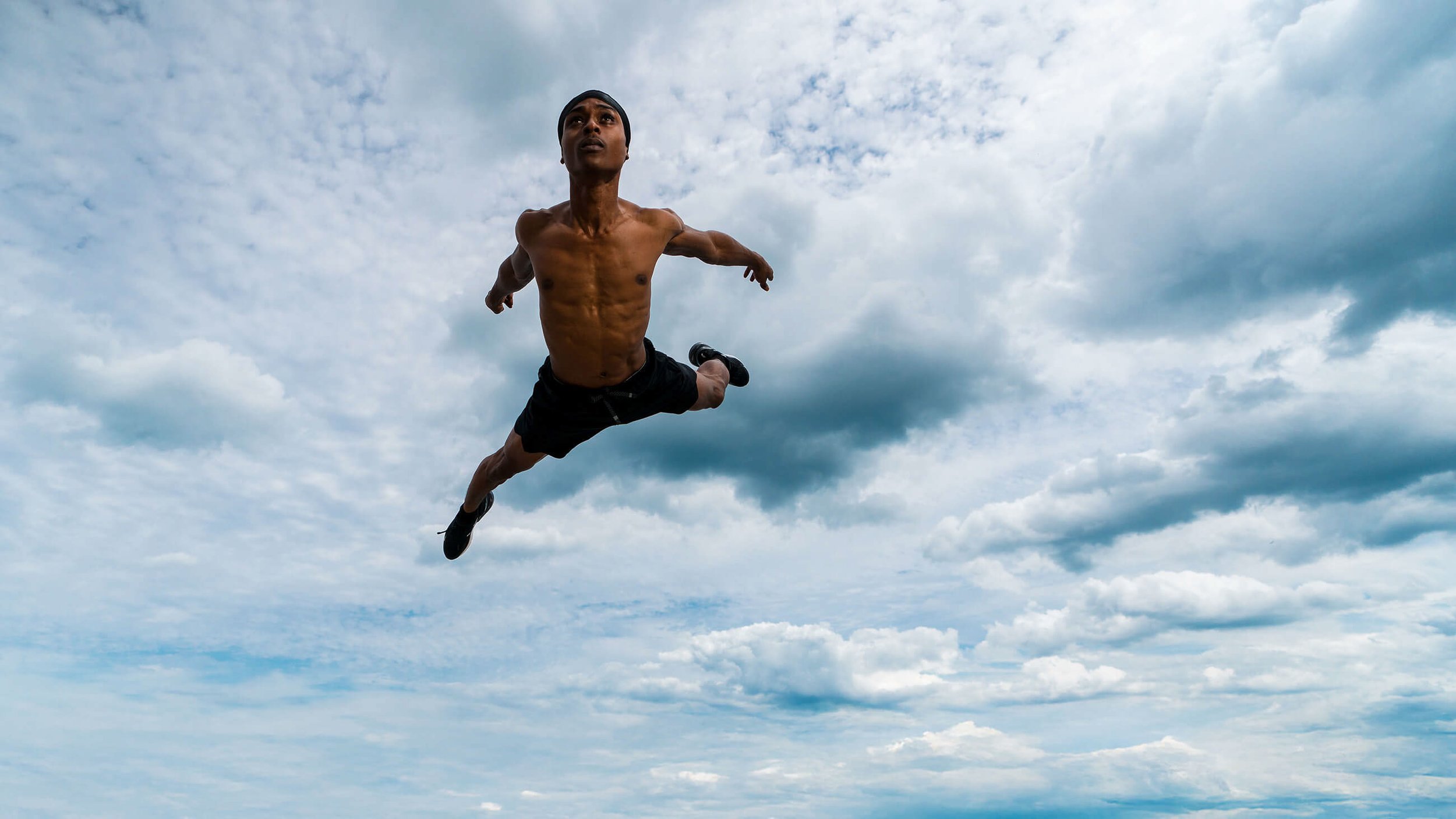 A shirtless man wearing black shorts and a headband leaps into the air against a partly cloudy sky, mid-flight.