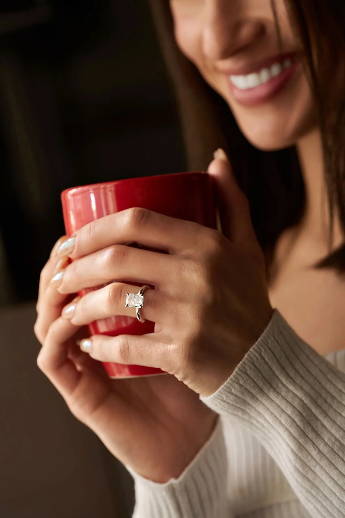 Close-up of a woman's hands holding a red mug, showing her engagement ring with a large square diamond.