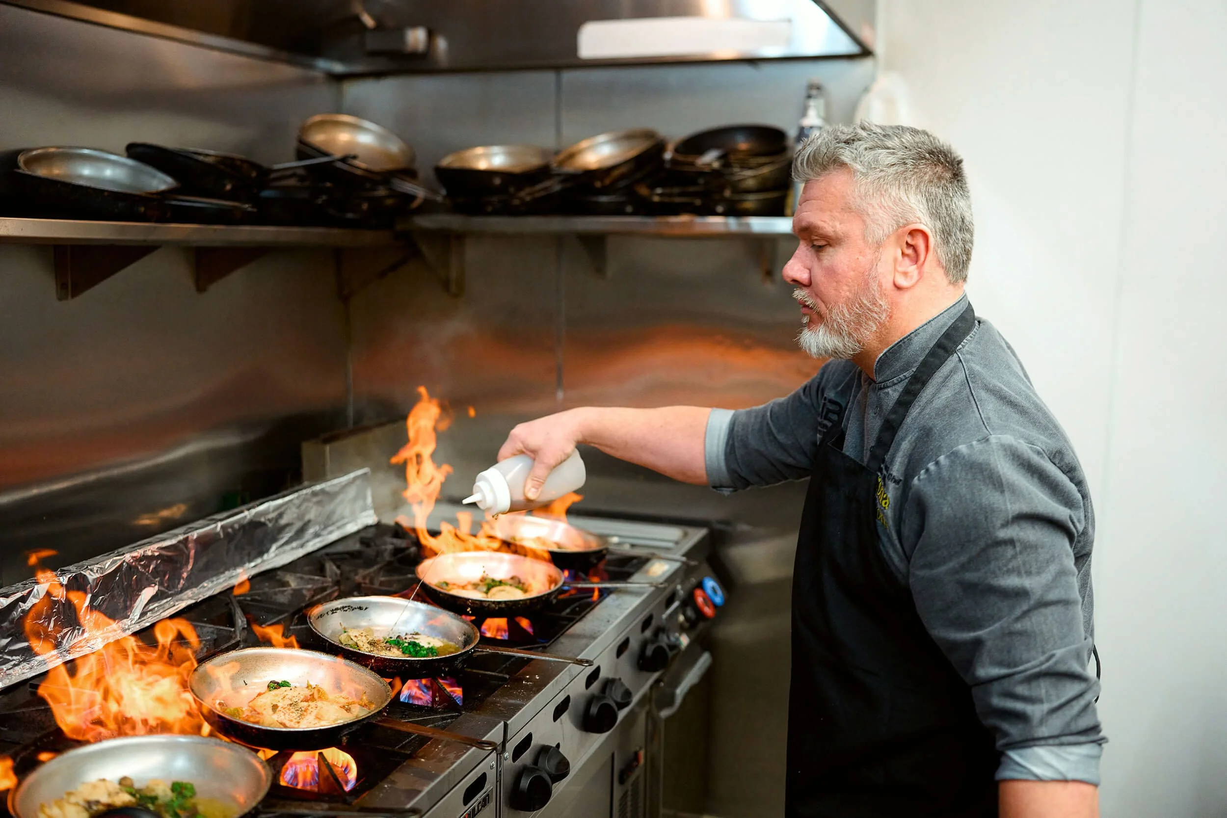 A chef in a gray shirt and black apron is cooking on a stove with multiple flames and pans. He is spraying something into a pan, causing a flare-up. There are stacked pans on a shelf above the stove.