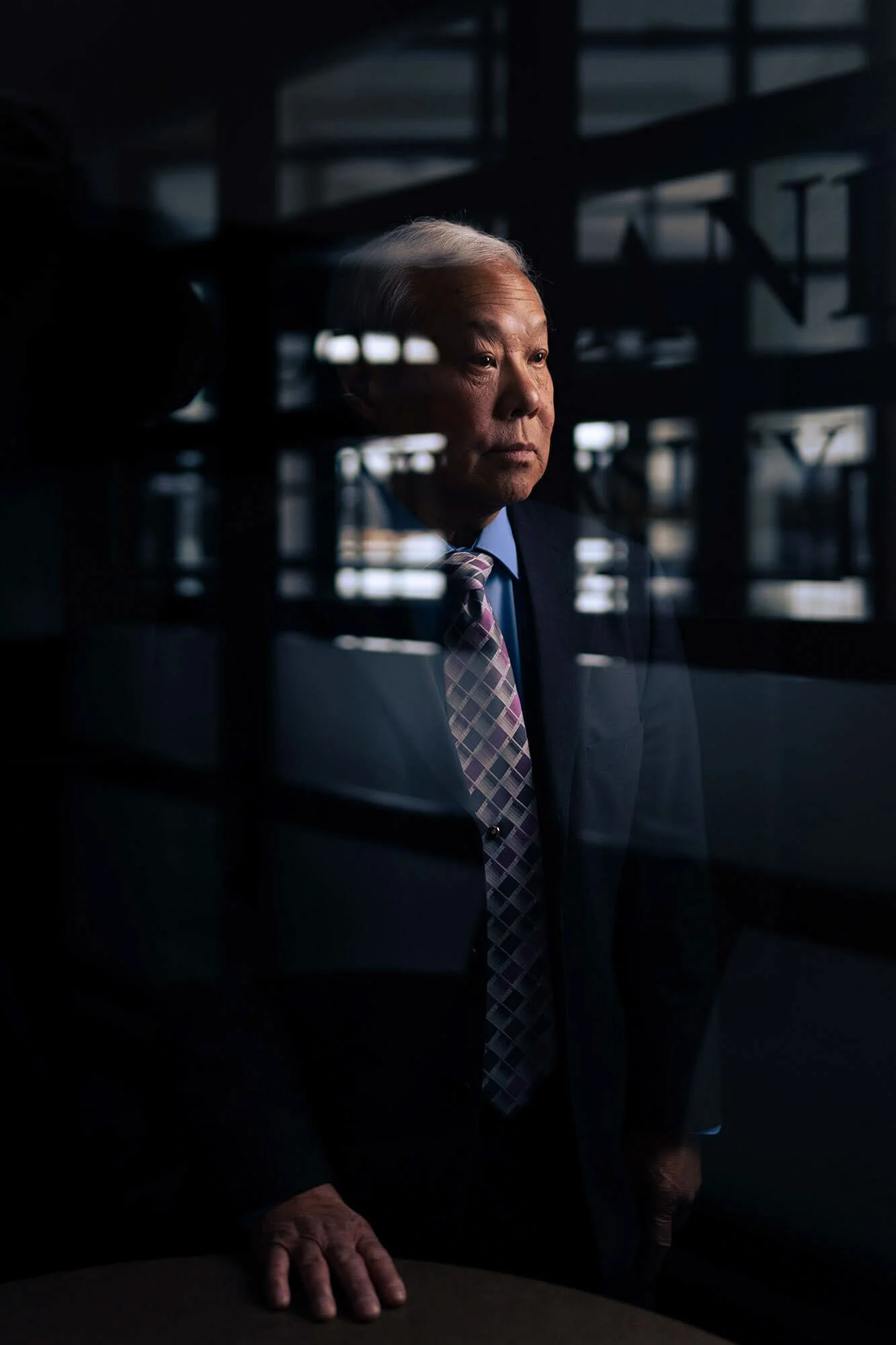 An elderly man with gray hair, dressed in a suit and patterned tie, standing in a dimly lit room, viewed through glass with reflections.