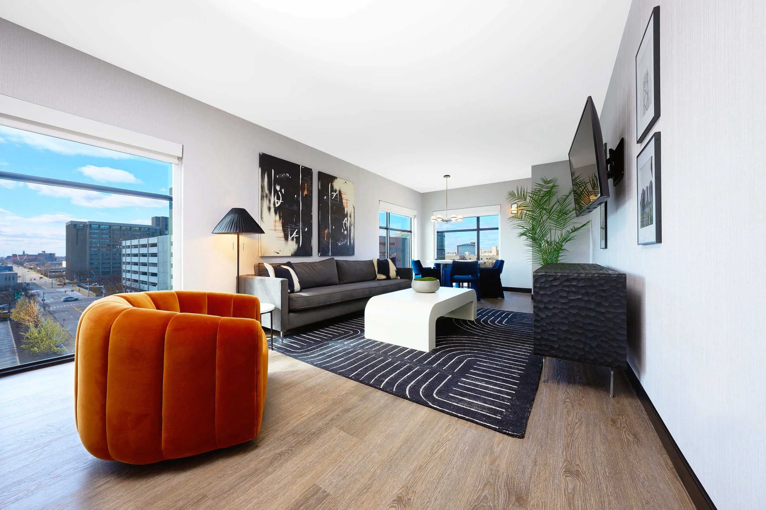Modern living room with large windows, gray sofa, orange armchair, white coffee table, black and white artwork, TV on wall, and potted plant.