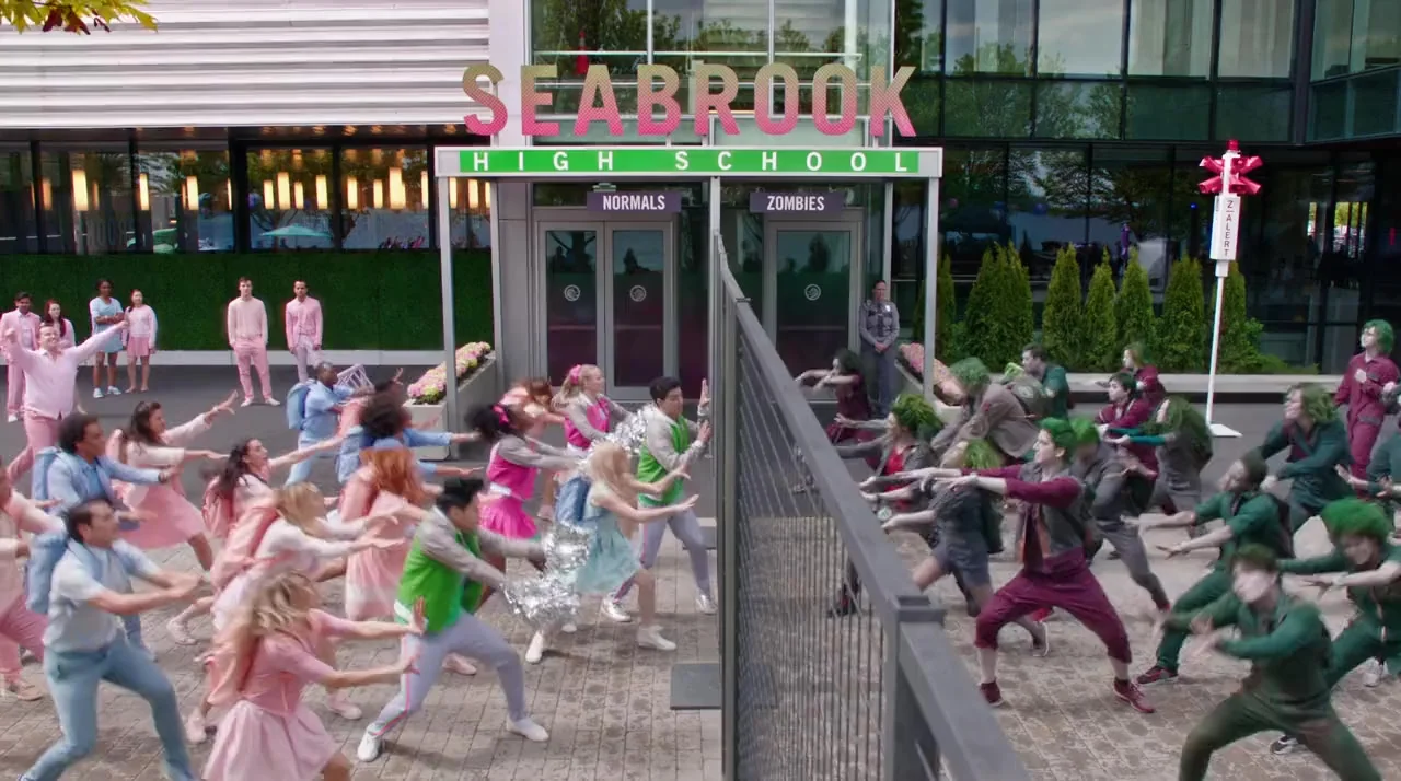 The central conflict of Disney's Z-O-M-B-I-E-S is the first day of school integration. Zombies are the hated underclass of Seabrook. Their homicidal cravings having been brought under control by an inhibiting device (the Z-Band), they are now just normal people. But Seabrook still imposes a curfew, offers only menial work and lives in fear of the Zombies. Here the pastel fearfulness of Seabrook is confronted by the saturated, exuberant excitement of the young zombies.