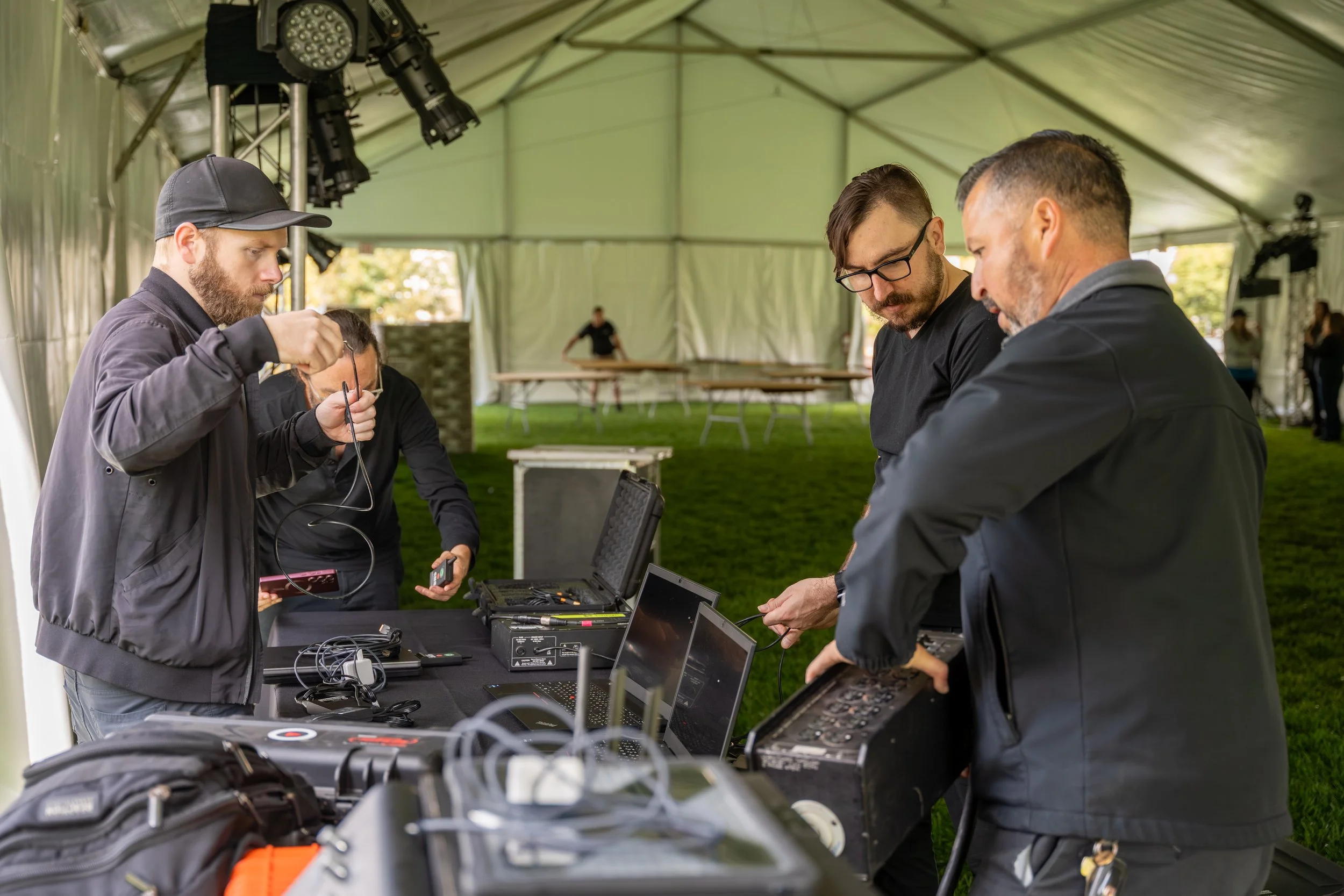 4 technicians set up AV equipment on a table in an event tent. Behind them are lighting truss and folding tables being assembled in the tent