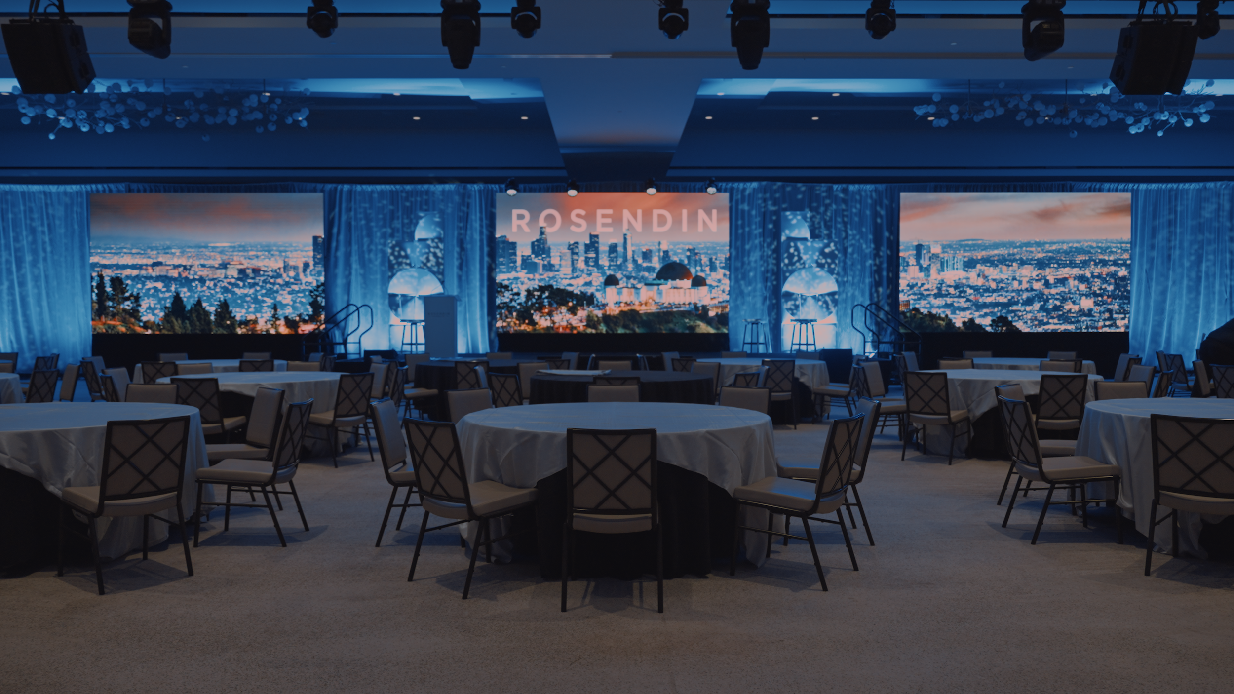 A room set for dinner, with draped round tables in front of a stage with 3 LED Walls. Lighting is visible at the top of the frame hanging from a truss.