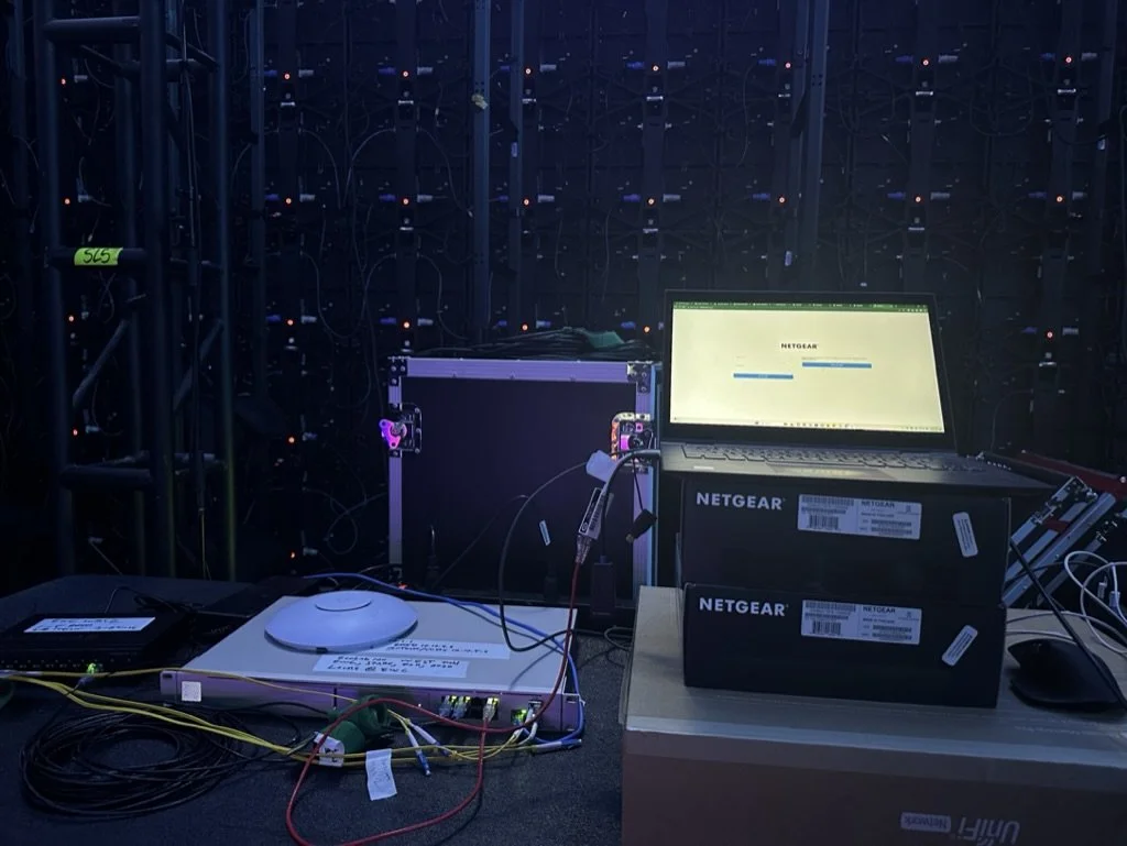 A laptop, a network switch, and an access point sit on a table backstage at an event. The table is facing the back side of an LED Wall.