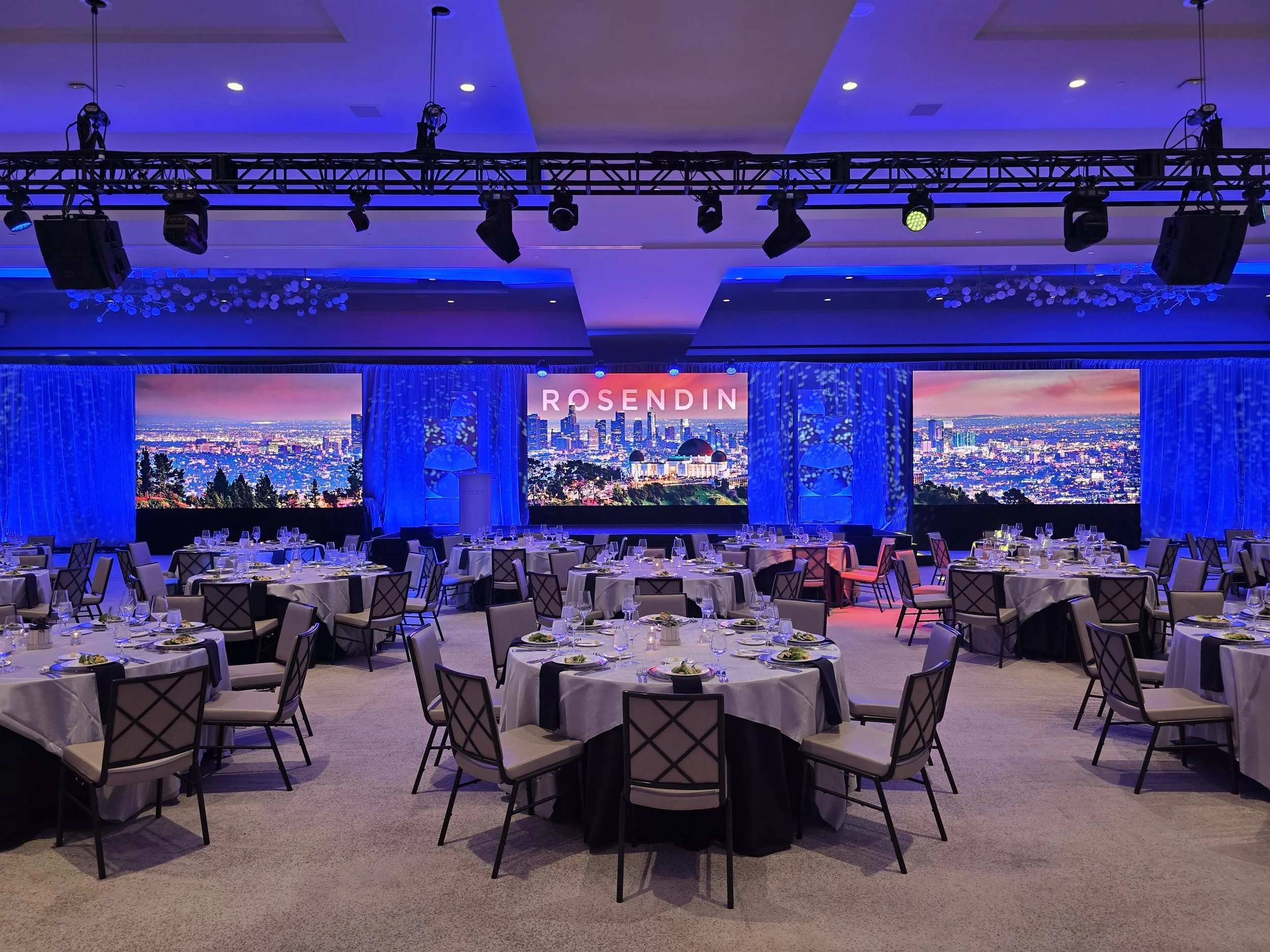 Three Screens sit in the front of a hotel ballroom with uplit drape and scenic visible. Above a truss holds lights and speakers, and tables are set for a banquet dinner.