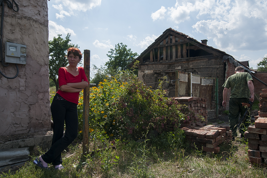 Woman standing next to her house as Aleksei walks towards the front line position hold by his men