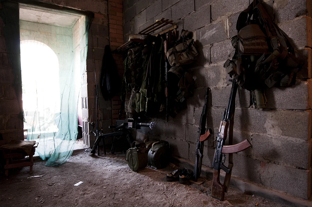 Fishing rob standing behind a grenade launcher and other military gear in the Patriot Battalion base located on the front line