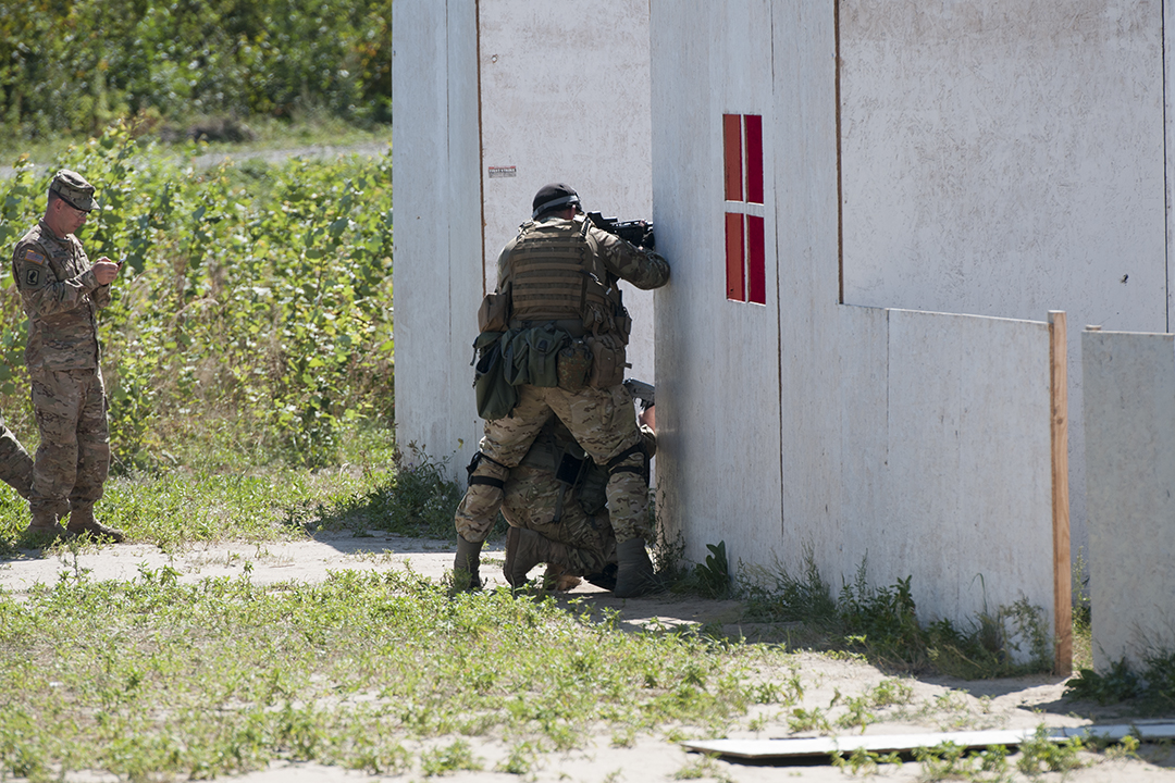 Ukrainian soldiers peaking at the corner of a fake building while an American officer monitor the exercise.