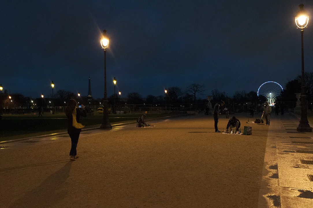 The unilluminated Eiffel Tower detaching itself from the landscape as a woman take pictures of street hawkers.