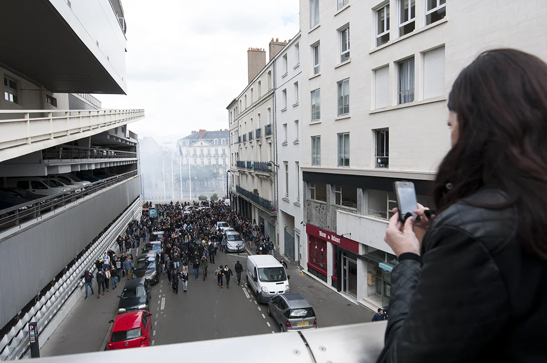 Young woman taking pictures with her cellphone of protesters engaging the rally away from tear gas fired at them by Police forces.