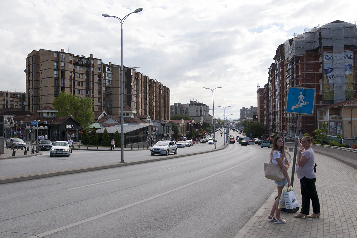 Tow women waiting for their bus on the sidewalk of a large boulevard.