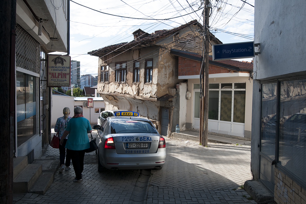 Two women walking past a taxi doing a u-turn in a small street of Pristina