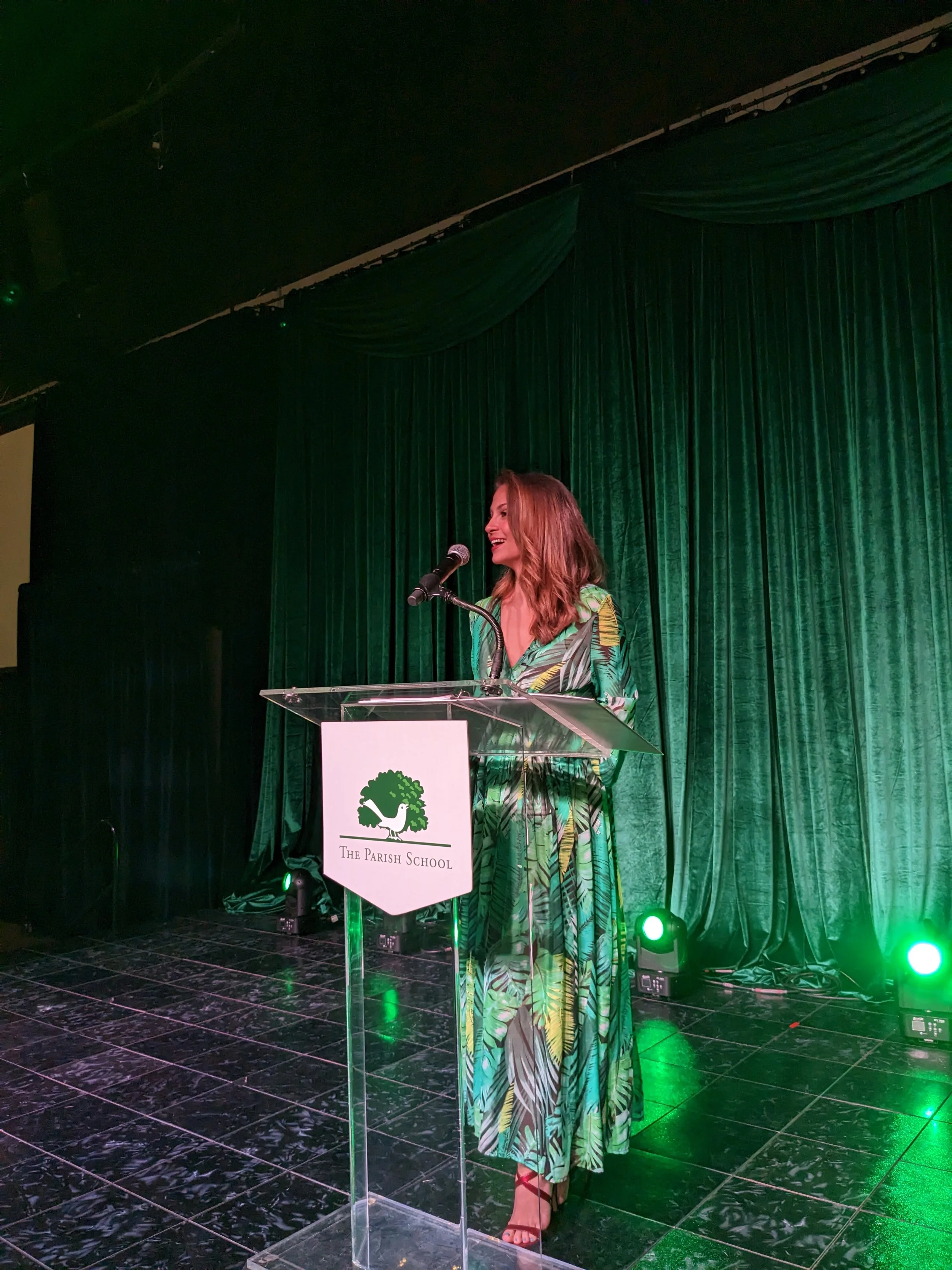 Kaitlin Monte stands at a podium in front of a green curtain. She speaks into a microphone while emceeing a non-profit fundraiser.