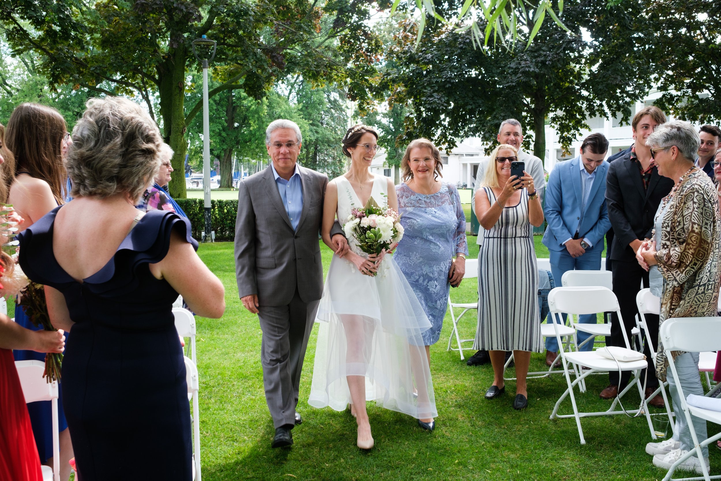  A colour wedding candin  photograph of Rocio’s parents walking her down the aisle during her and Geoff’s outdoor wedding ceremony at the Royal Canadian Yacht Club by Toronto wedding photographer Scott Williams  