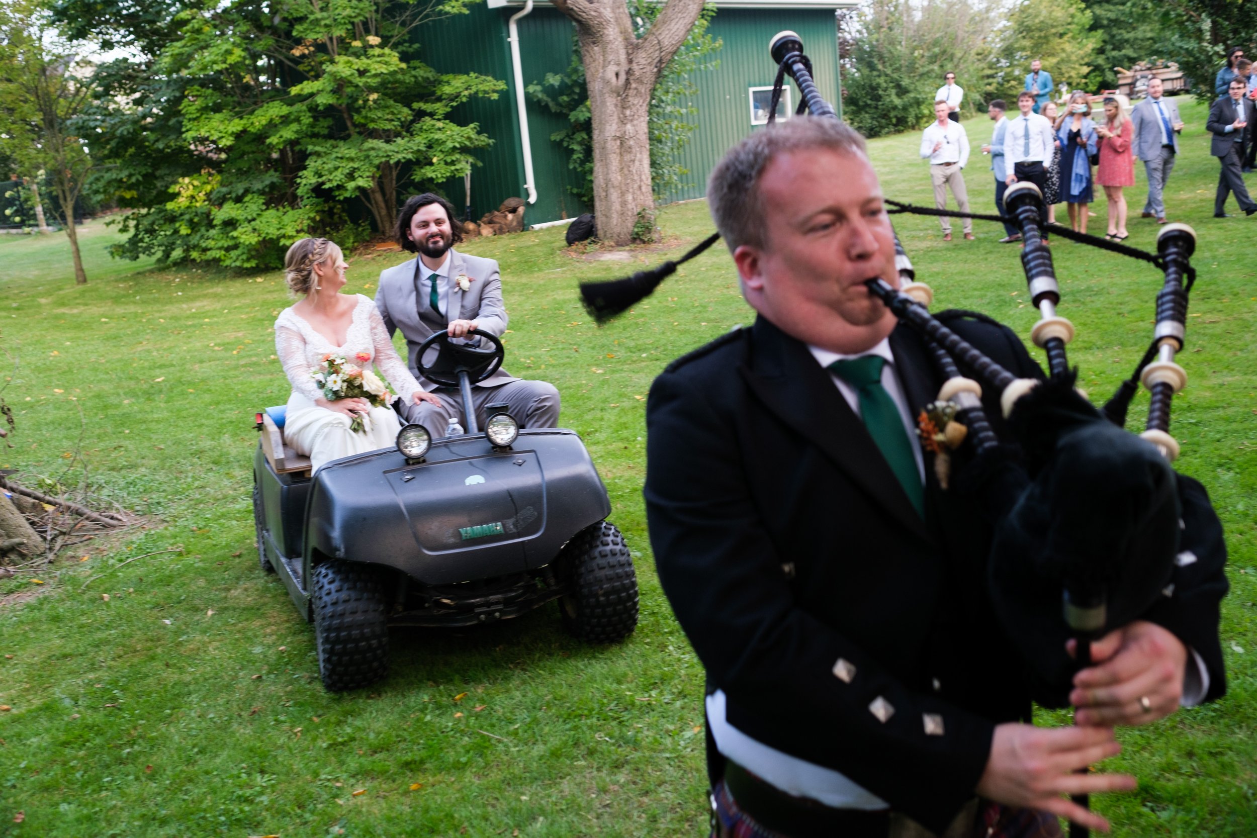  A colour candid wedding photograph of the bride and groom entering their cocktail hour with a bagpiper during Ailish + Evan’s outdoor farm wedding near Pickering, Ontario by Toronto wedding photographer Scott Williams (www.scottwiliiamsphotographer.
