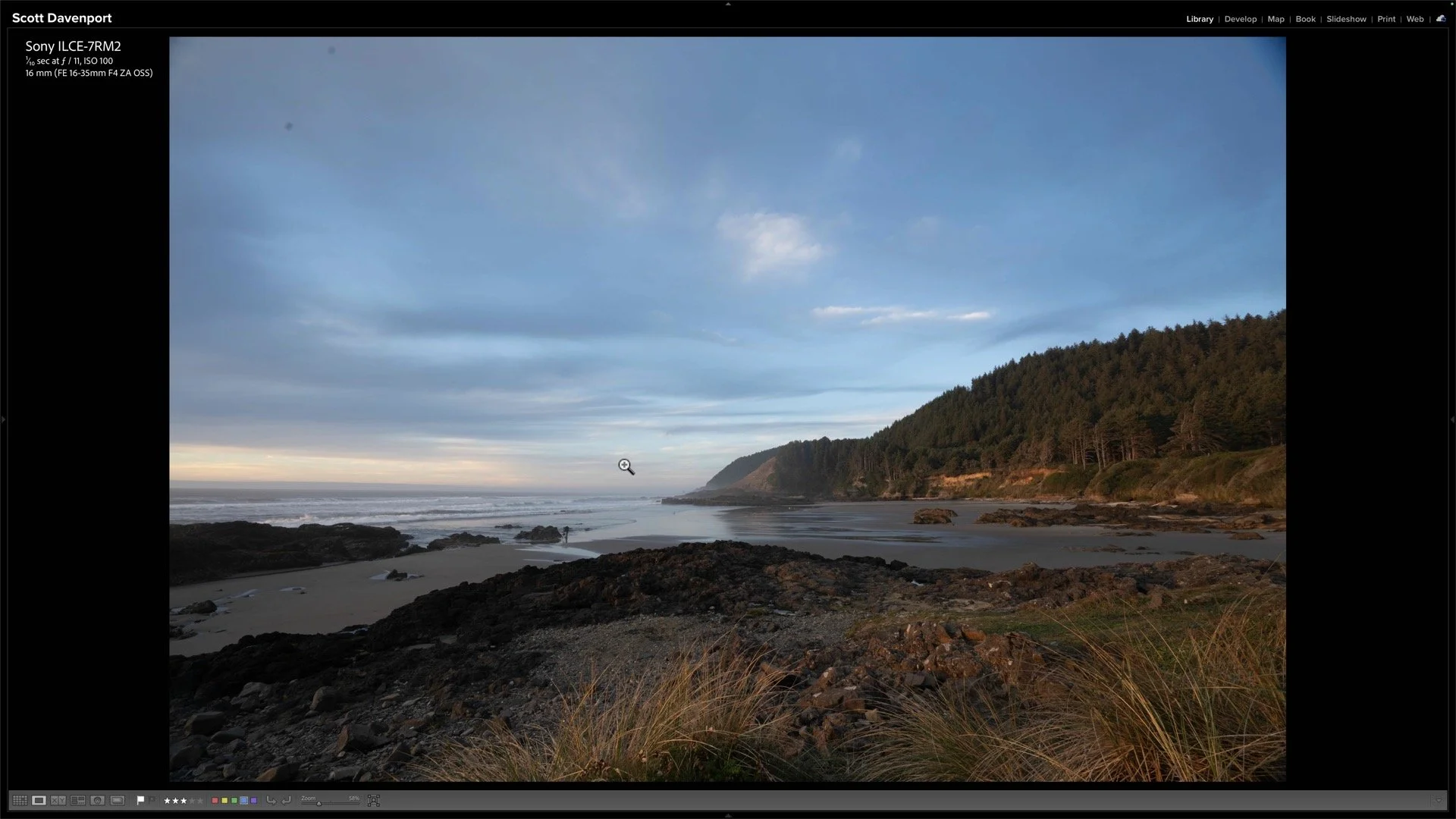 Golden sunlight catches grasses above a reflective beach, with coastal headlands and a colorful sky beyond.