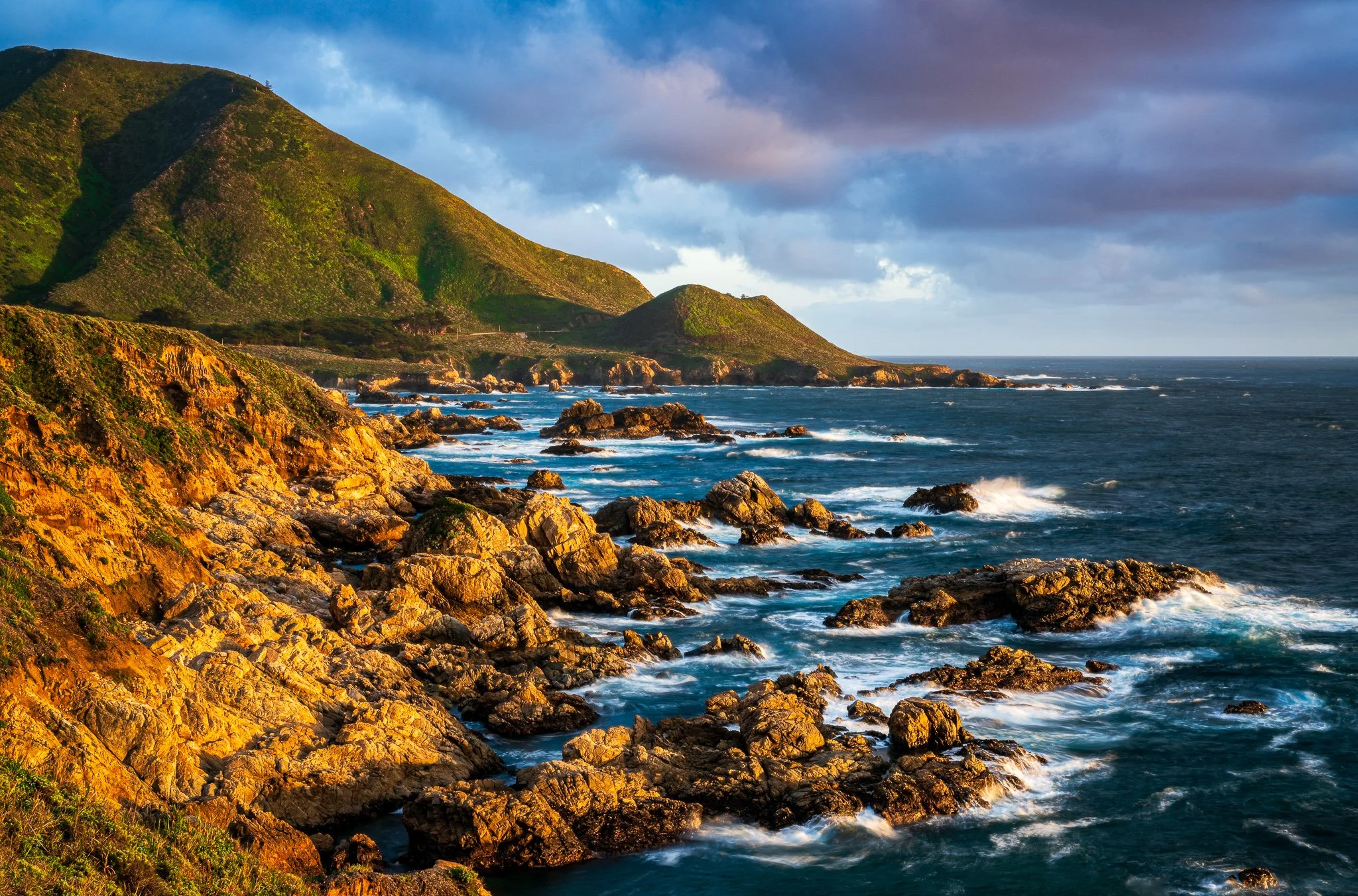 Warm evening light crosses a rugged shoreline at Soberanes Point, with rocky coves leading toward green coastal hills beneath blue-gray clouds.