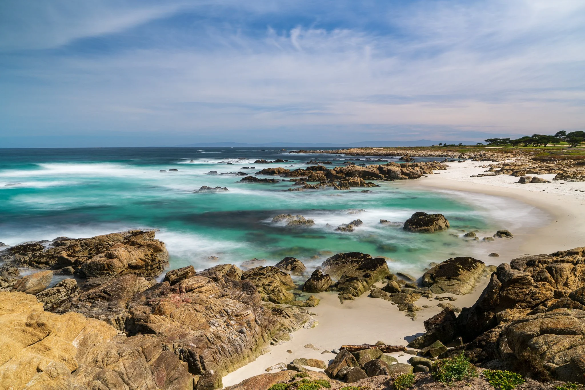 White sand, granite outcrops, and turquoise water curve through Granite Beach beneath streaked late-morning clouds.