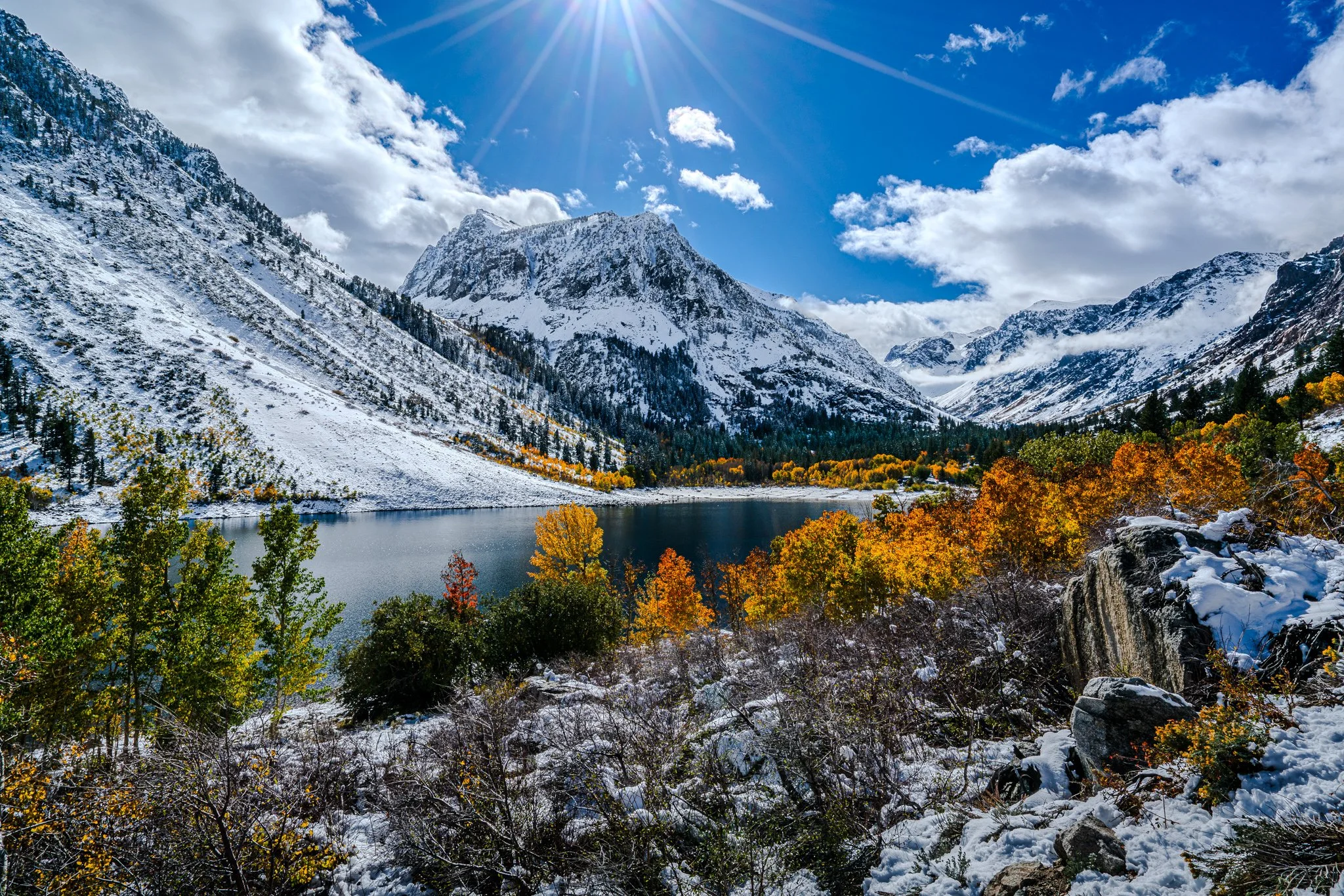 Lundy Lake sits beneath freshly snow-covered canyon walls, with golden autumn foliage along the shore and sunlight breaking through clouds.