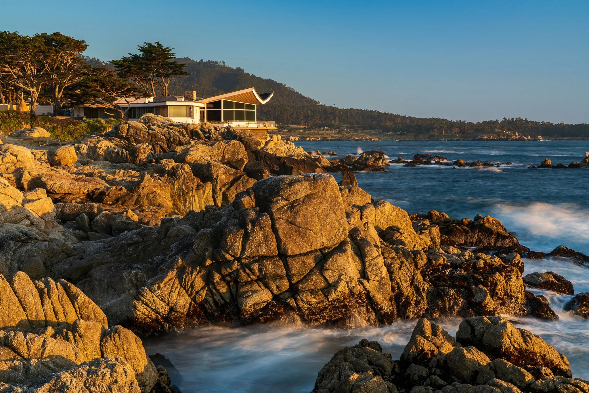 Modern coastal house above sunlit granite rocks at Carmel Point, with waves flowing around the shoreline beneath a clear blue sky.