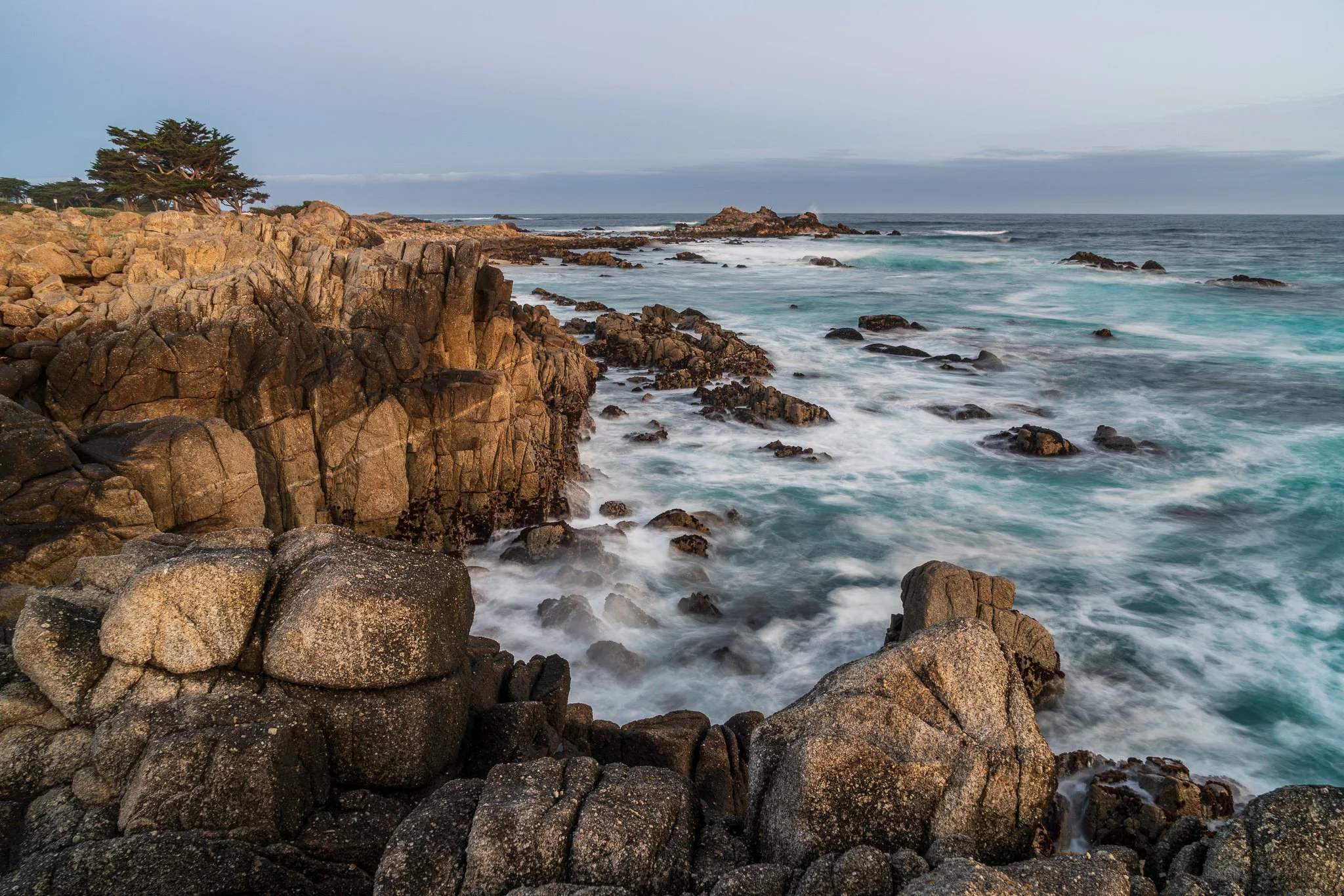 Granite ledges frame pale turquoise water along the Monterey Peninsula beneath a soft gray-blue dawn sky.