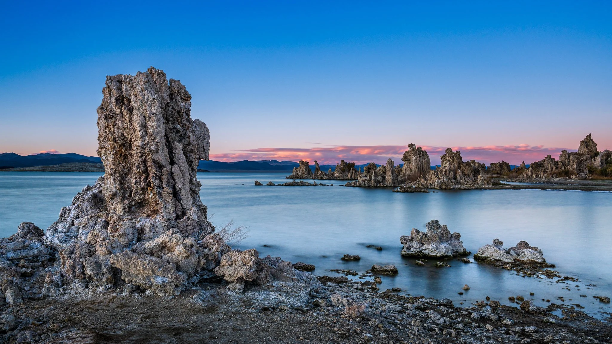 A tall tufa tower stands near the Mono Lake shoreline against calm blue water and a pink twilight sky..
