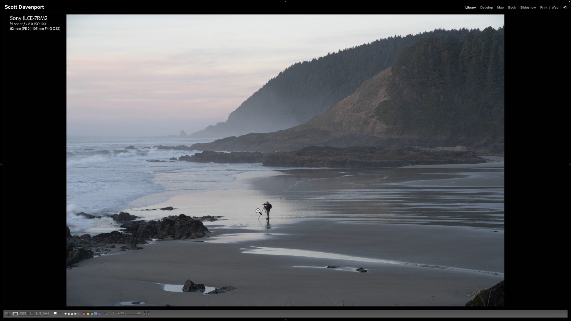 A lone photographer stands against a bright coastal background, framed by foreground rocks beneath misty headlands.