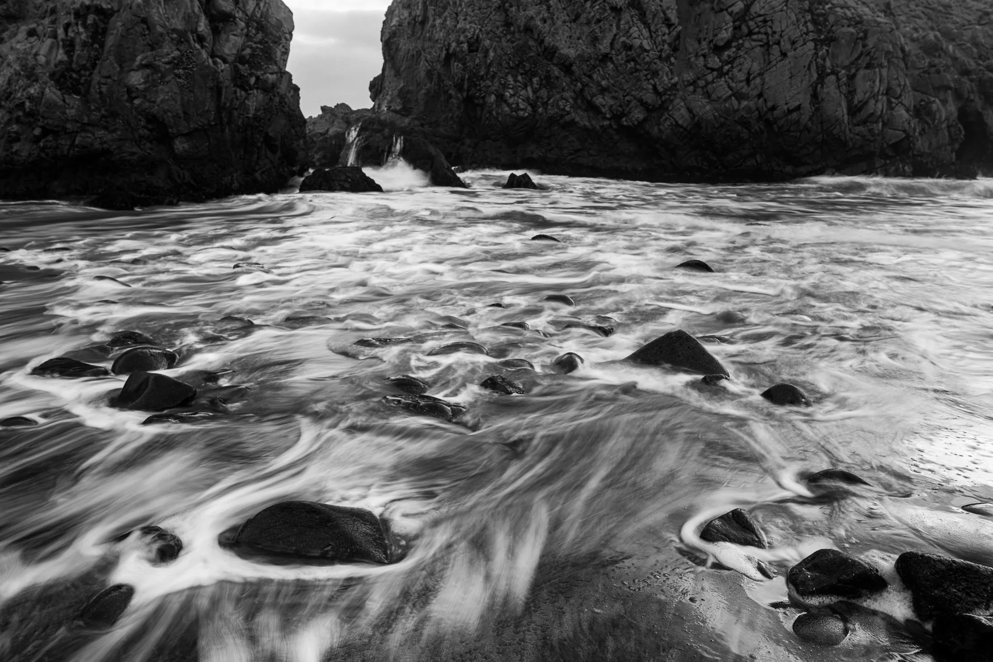 Foamy surf streams between dark rocks and through a large sea arch at Pfeiffer Beach in a black-and-white scene.