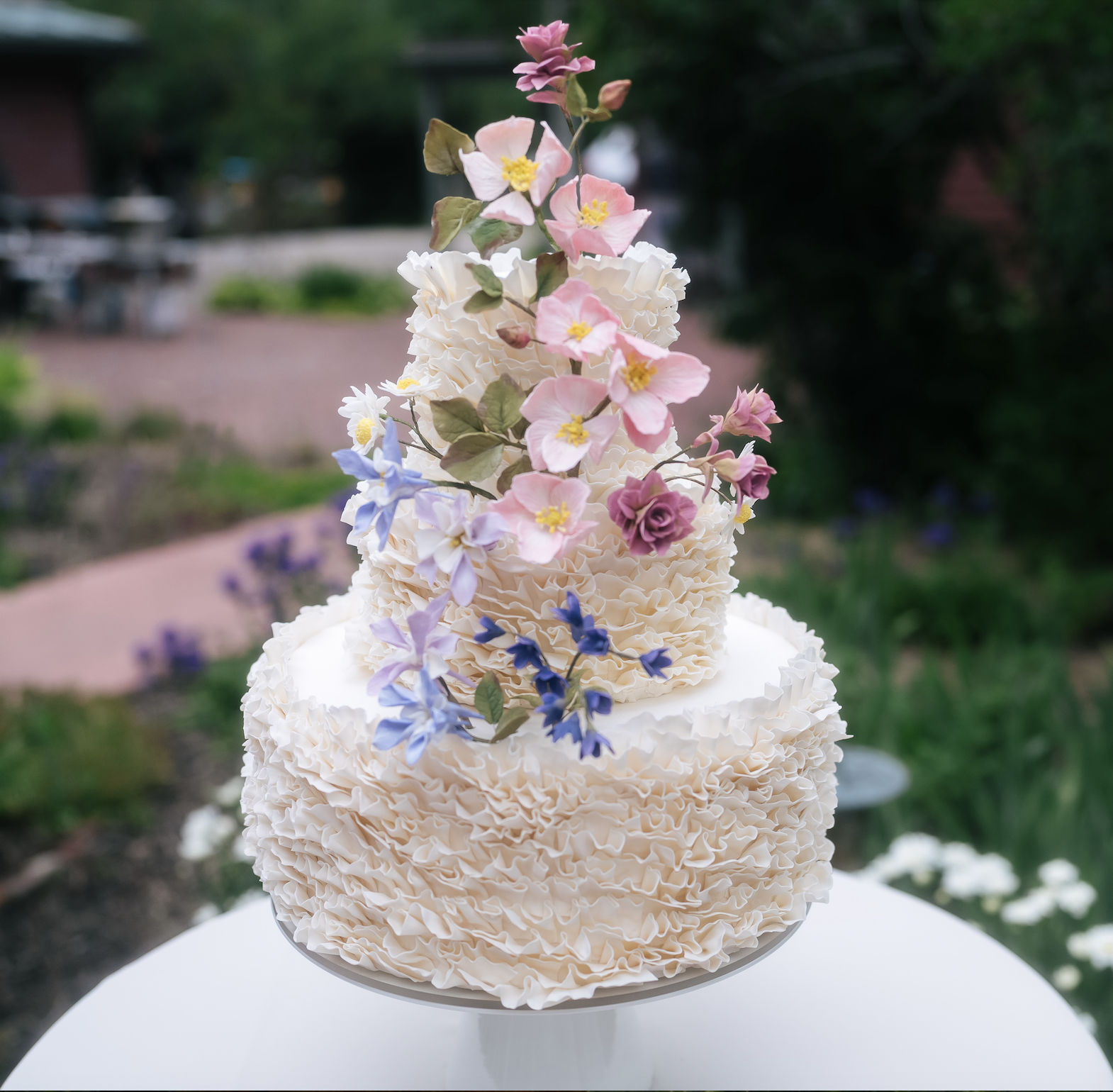 Two-tier wedding cake with ruffled white icing and pastel-colored flowers on top, outdoors in a garden setting.