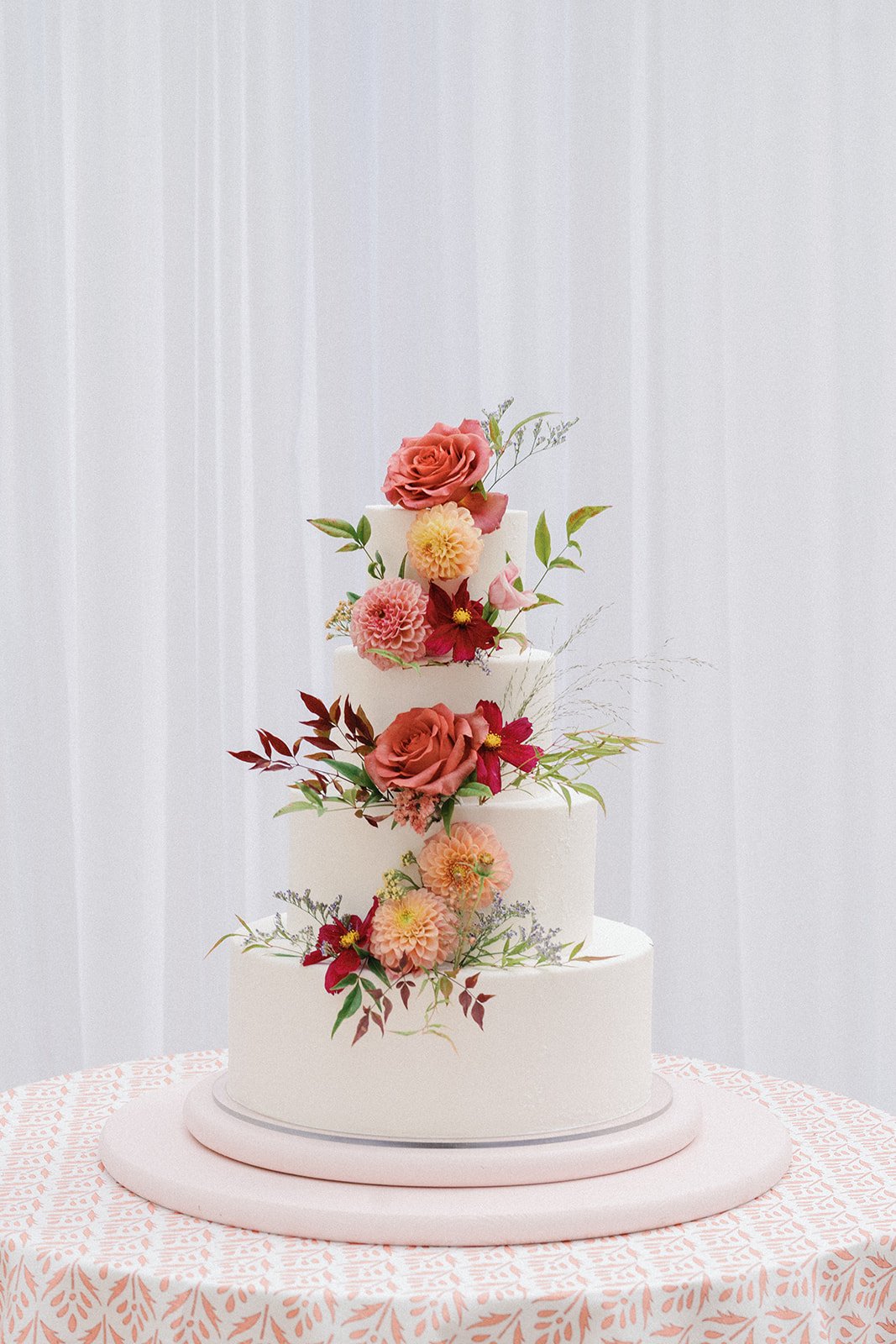 Three-tiered white wedding cake decorated with pink, red, and yellow flowers and green leaves, placed on a round table with a patterned pink tablecloth.