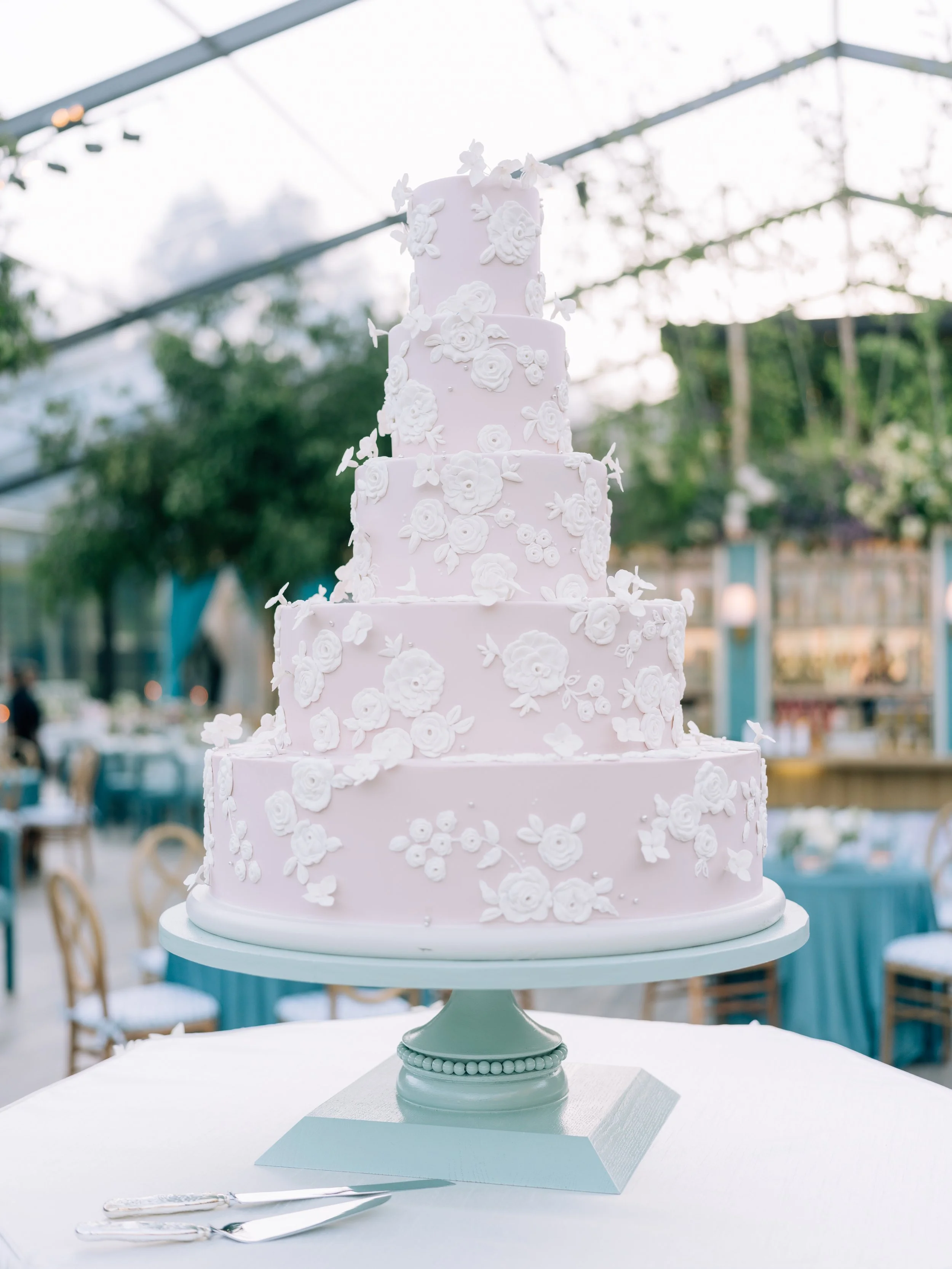 A tall, pink, five-tiered wedding cake decorated with white floral fondant flowers, displayed on a white cake stand at an outdoor event.
