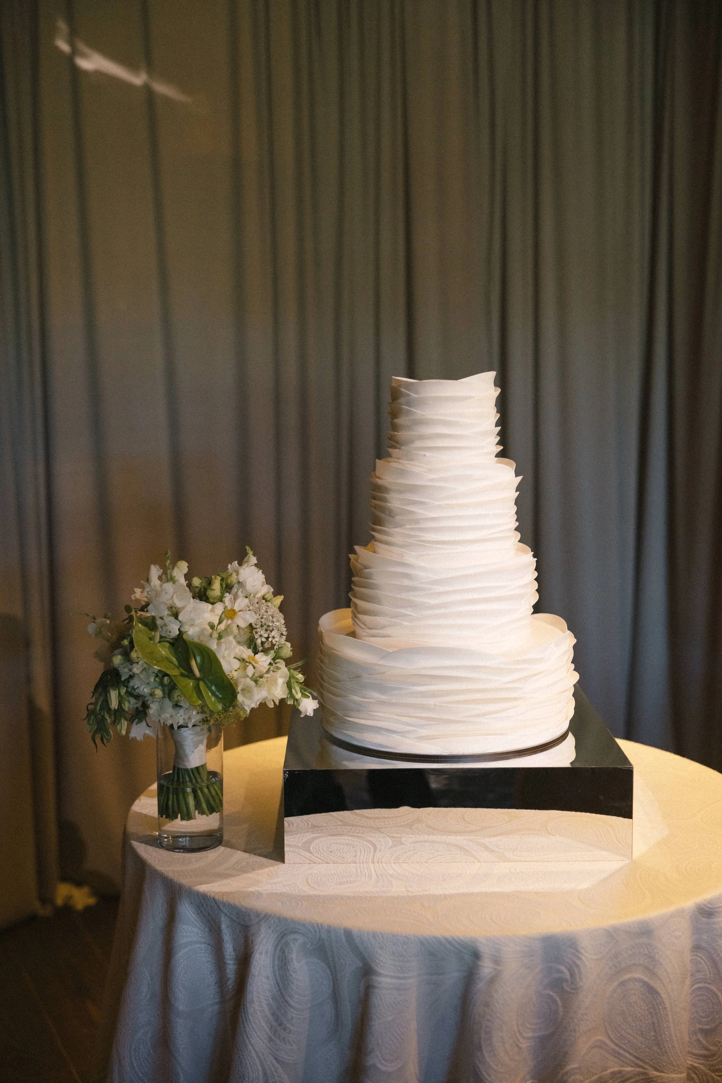 A four-tiered white wedding cake with textured frosting, on a reflective black base, next to a bouquet of white flowers in a glass vase, on a round table with a cream-colored tablecloth.
