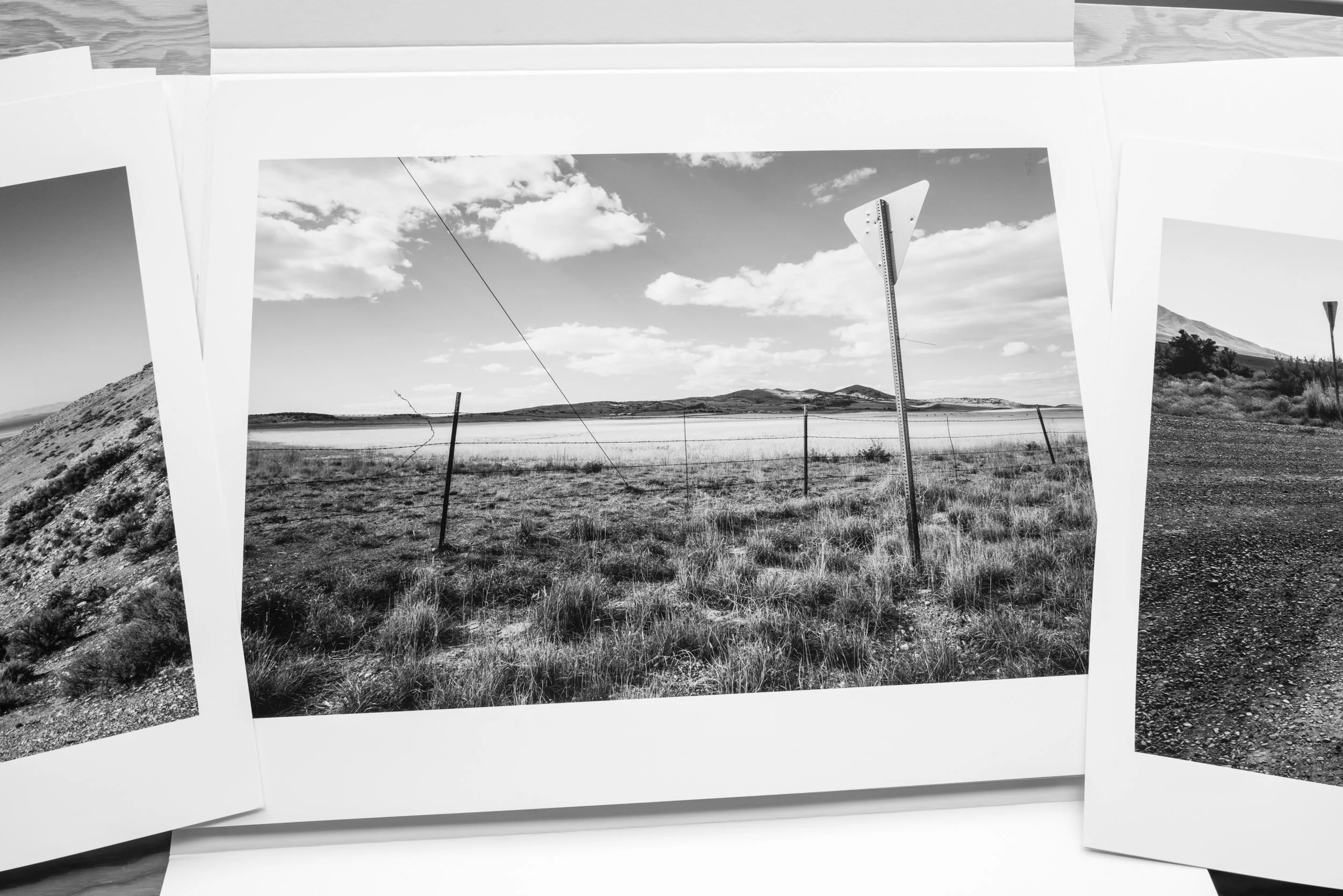 Black and white photograph of a landscape with mountains, a sky with clouds, a fence, and a road sign, framed within a white border and multiple photographic prints layered around it.