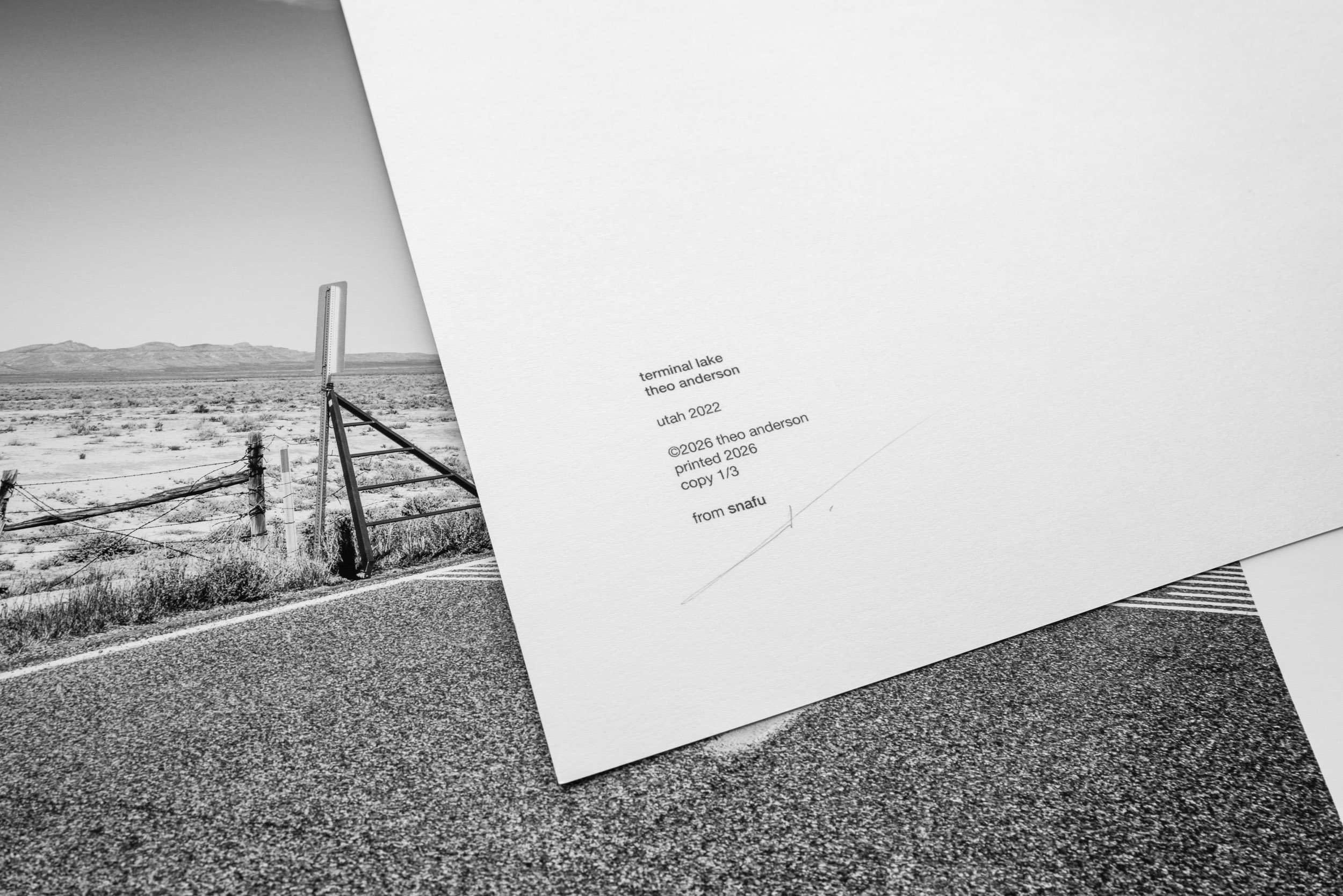 Black and white photograph featuring a desert landscape with a road, fence, and mountains in the background. Partially visible white paper with printed text overlays the photograph.