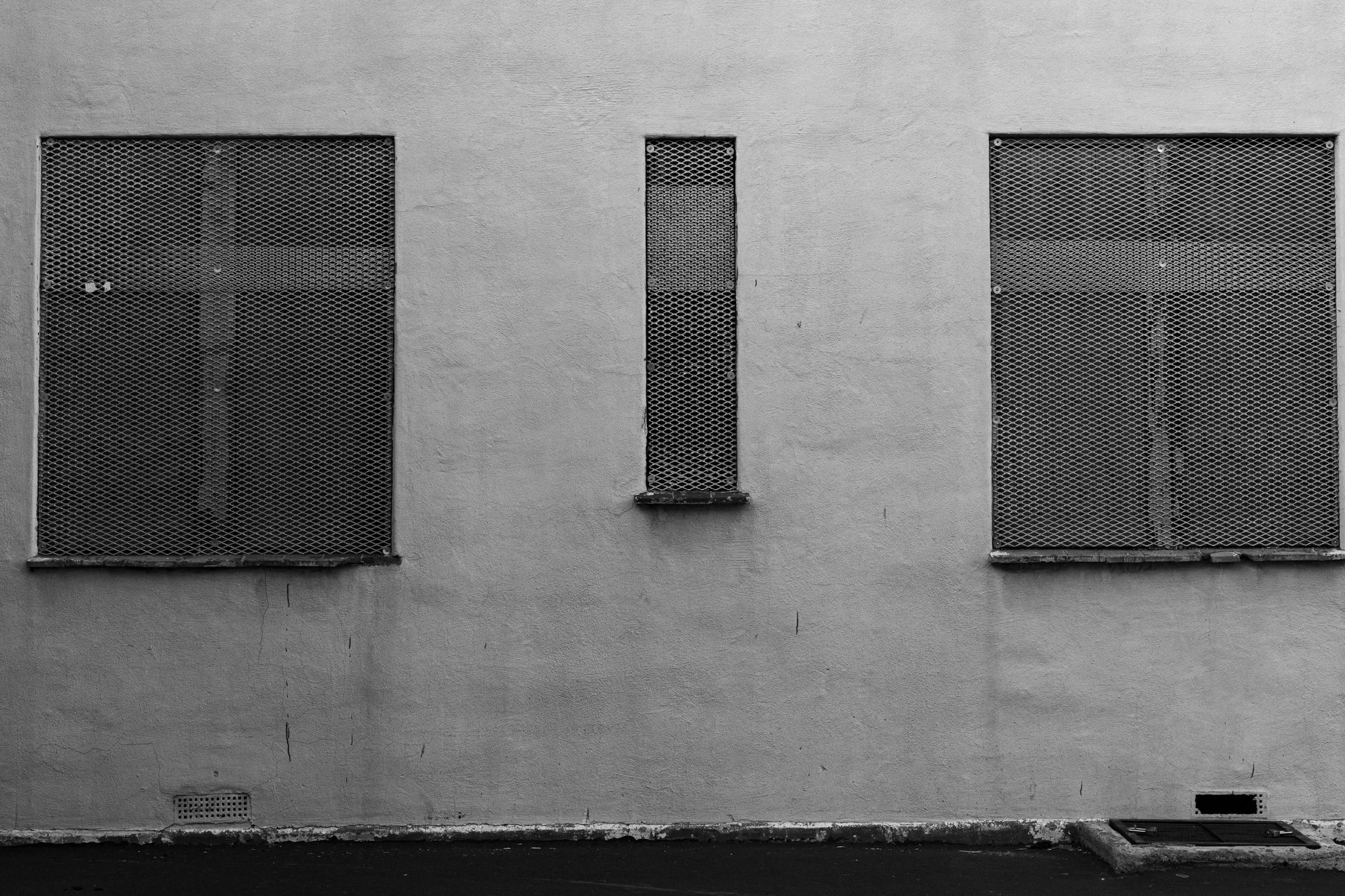 A black and white photograph of three windows covered in metal mesh grates on a light, textured wall. The composition is symmetrical, with two large rectangular windows flanking a narrow vertical one.