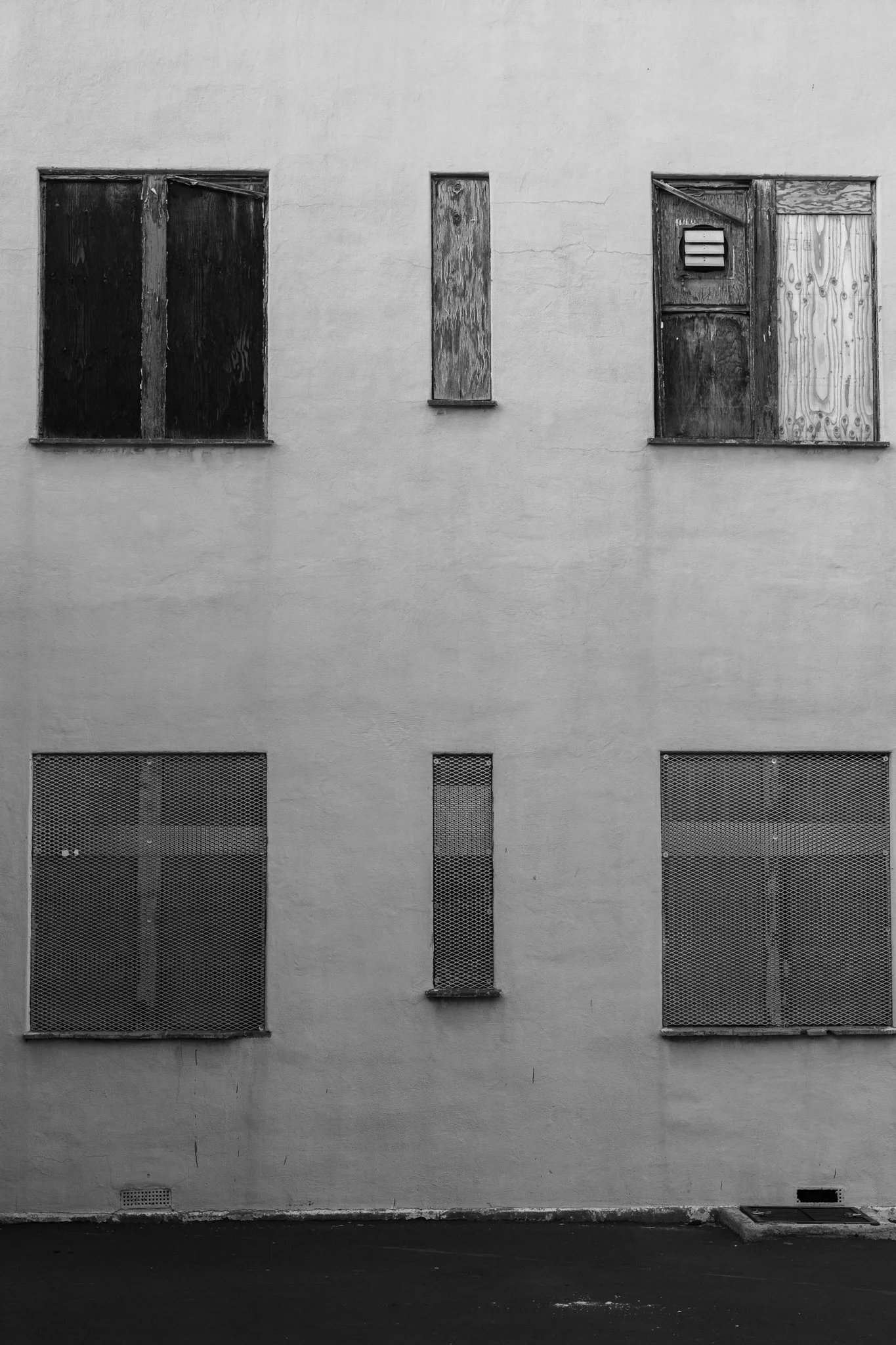 A vertical black and white shot of a building exterior featuring two tiers of windows. The bottom row of windows is covered with metal mesh, while the top row is boarded up with weathered plywood, creating a stark, urban contrast.