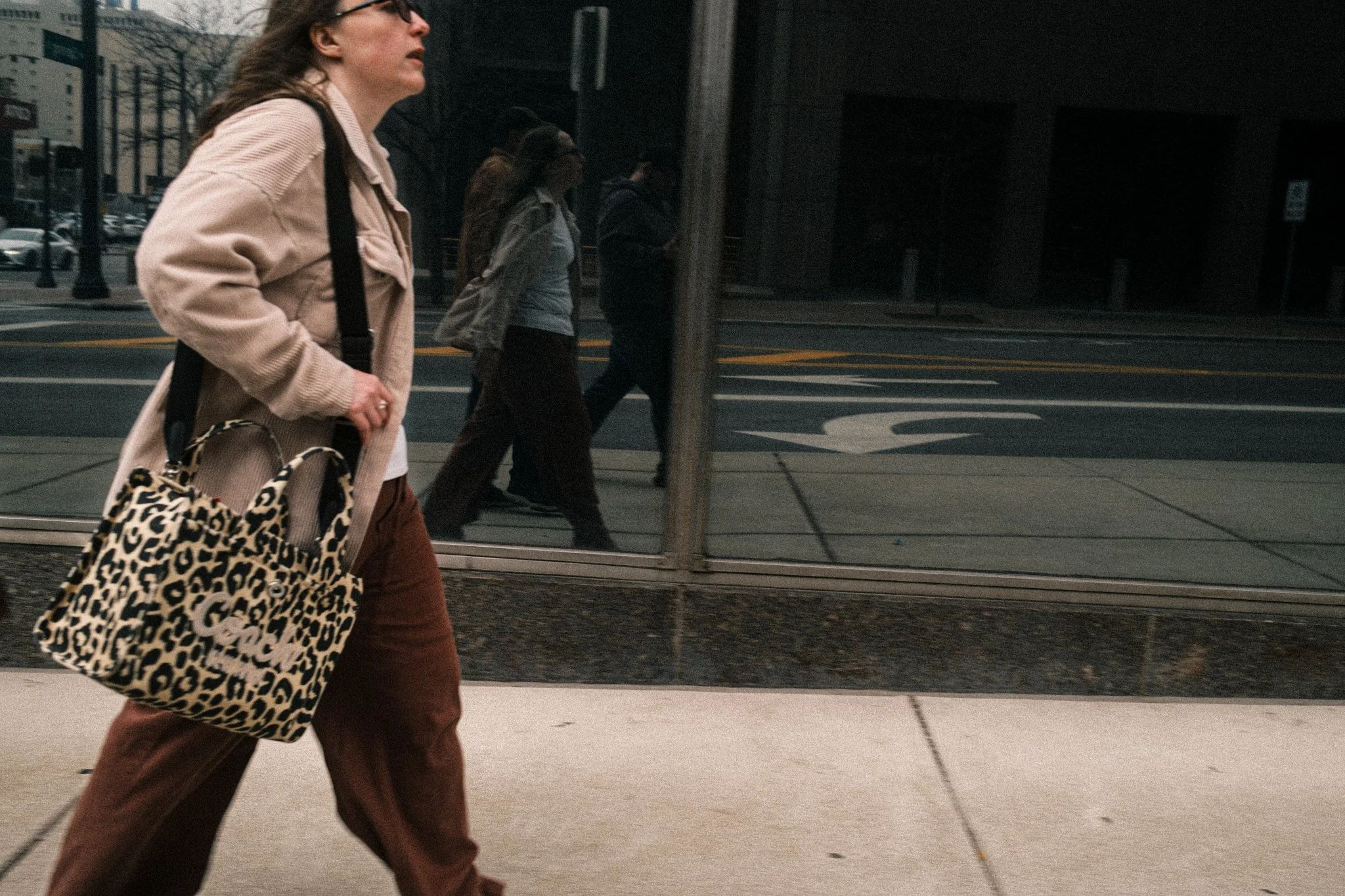 A woman walks briskly along a city sidewalk, her figure captured in a candid, street-photography style.
