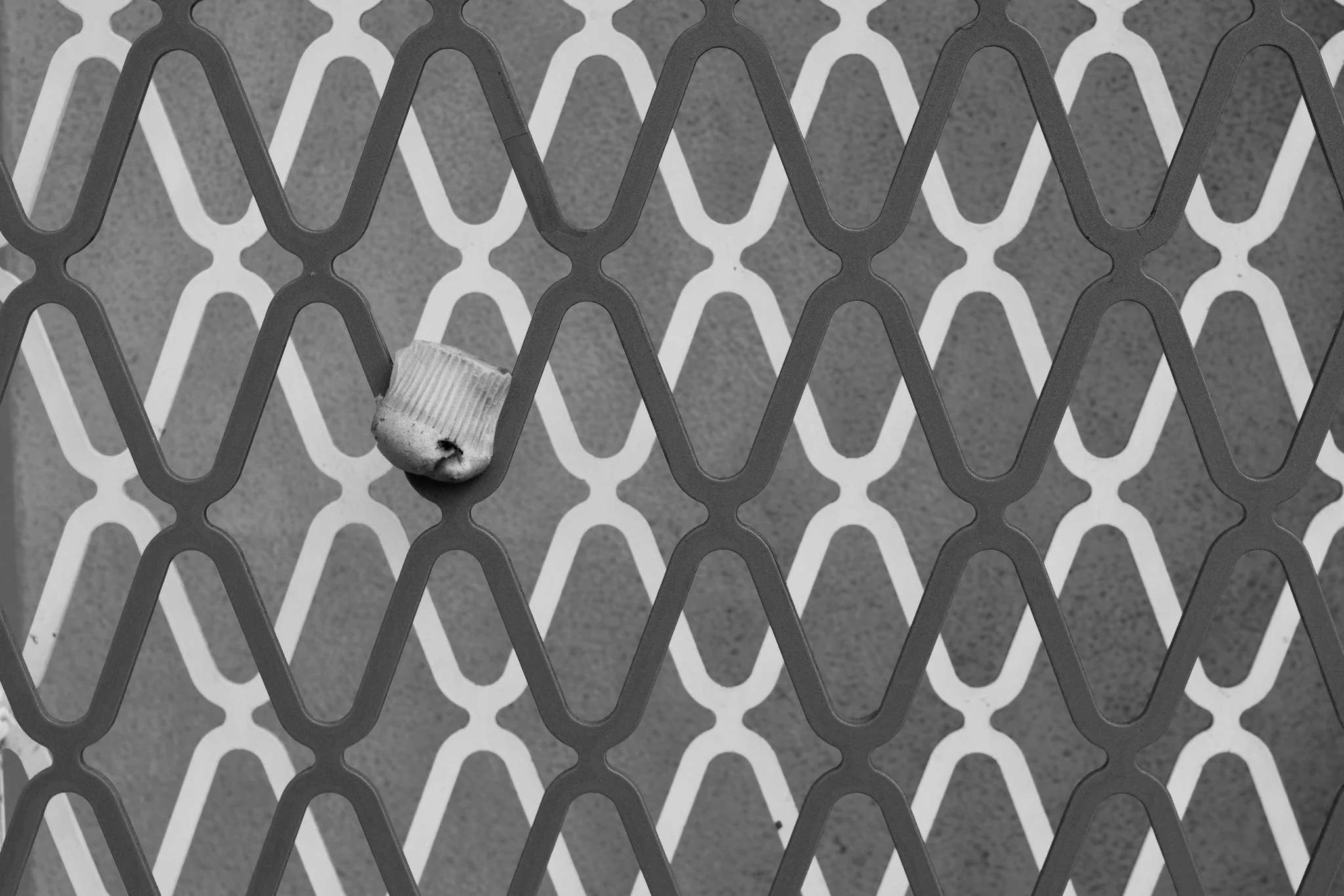 A black-and-white, close-up photograph featuring a repetitive geometric pattern of a metal grate. A discarded blueberry muffin is shoved upside down into one of the grates, breaking the symmetry.