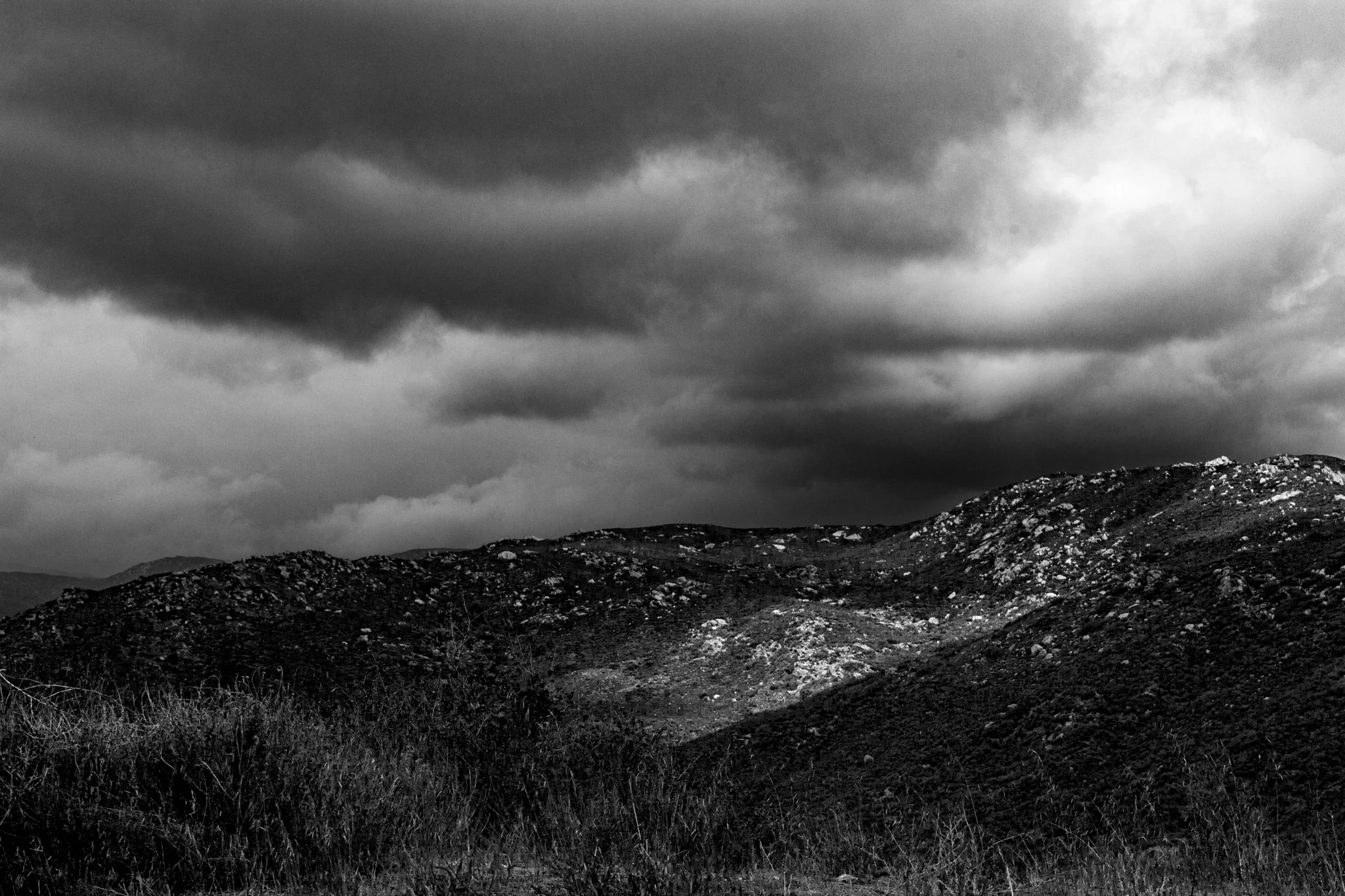 A dramatic black and white photograph of rolling, rocky hills under a heavy, storm-laden sky.