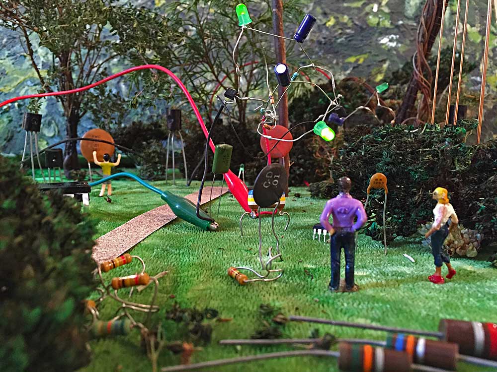A couple look at the green light emitted by an astable multivibrator living in the Electronic Sculpture Garden. In the background, a child plays with a ceramic capacitor while two transistors look on. Kelly Heaton, November 2015