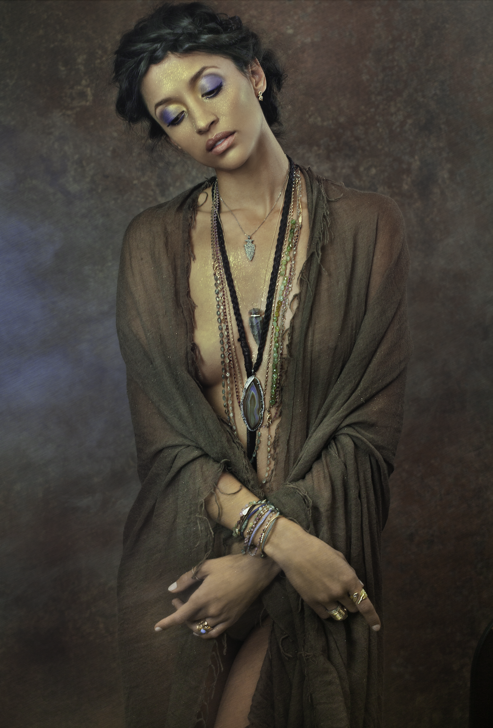 A woman with dark hair styled in braids, wearing purple and gold eye makeup, dressed in a tattered brown garment, and adorned with multiple layered necklaces, rings, and bracelets.