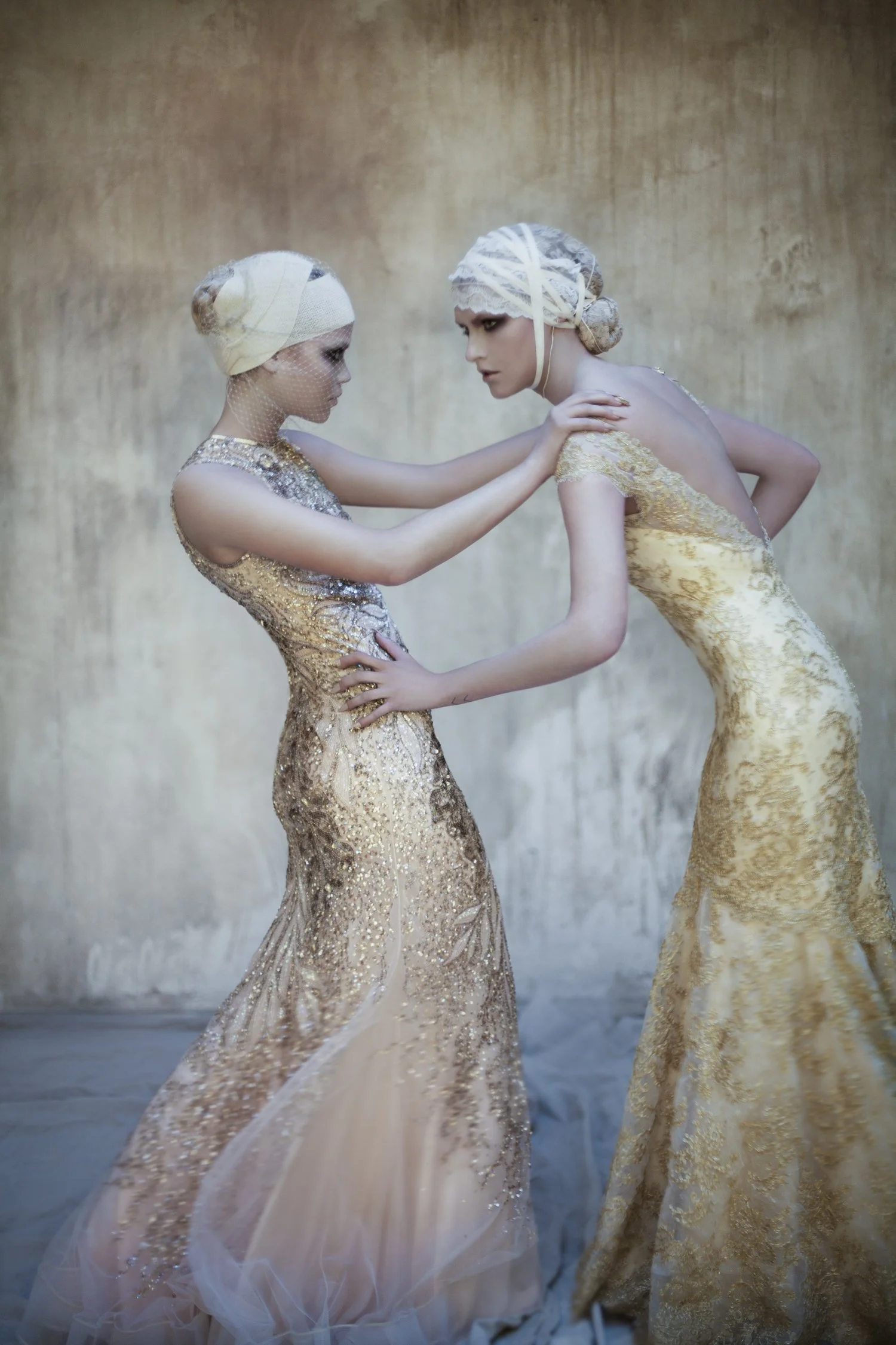 Two women in vintage style gold dresses and lace head coverings holding each other and staring into each other's eyes against a textured background.
