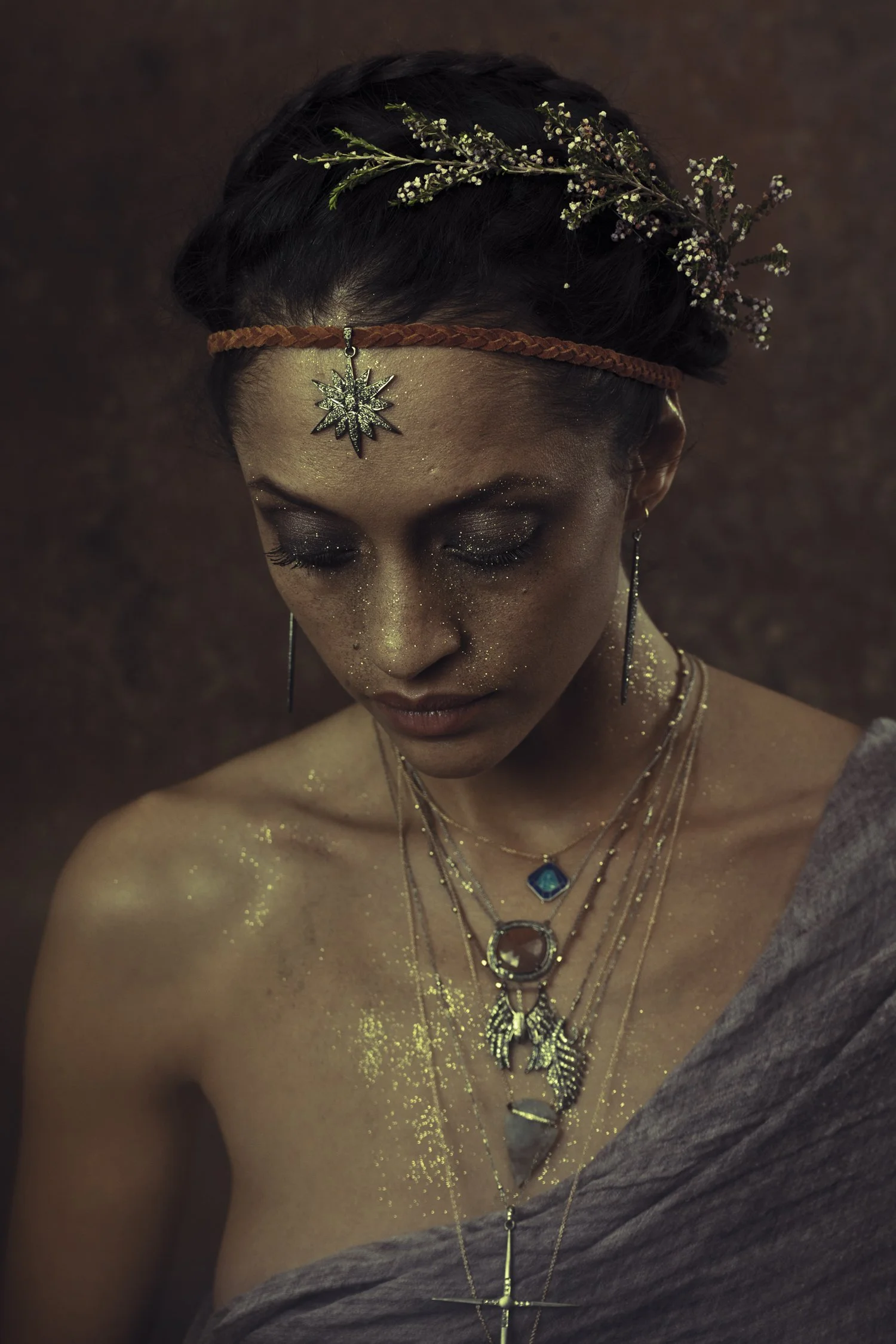 A woman with dark hair decorated with a floral crown and wearing layered necklaces with pendants, gold glitter on her face and shoulders, and a brown headband with a star-shaped pendant hanging from it.