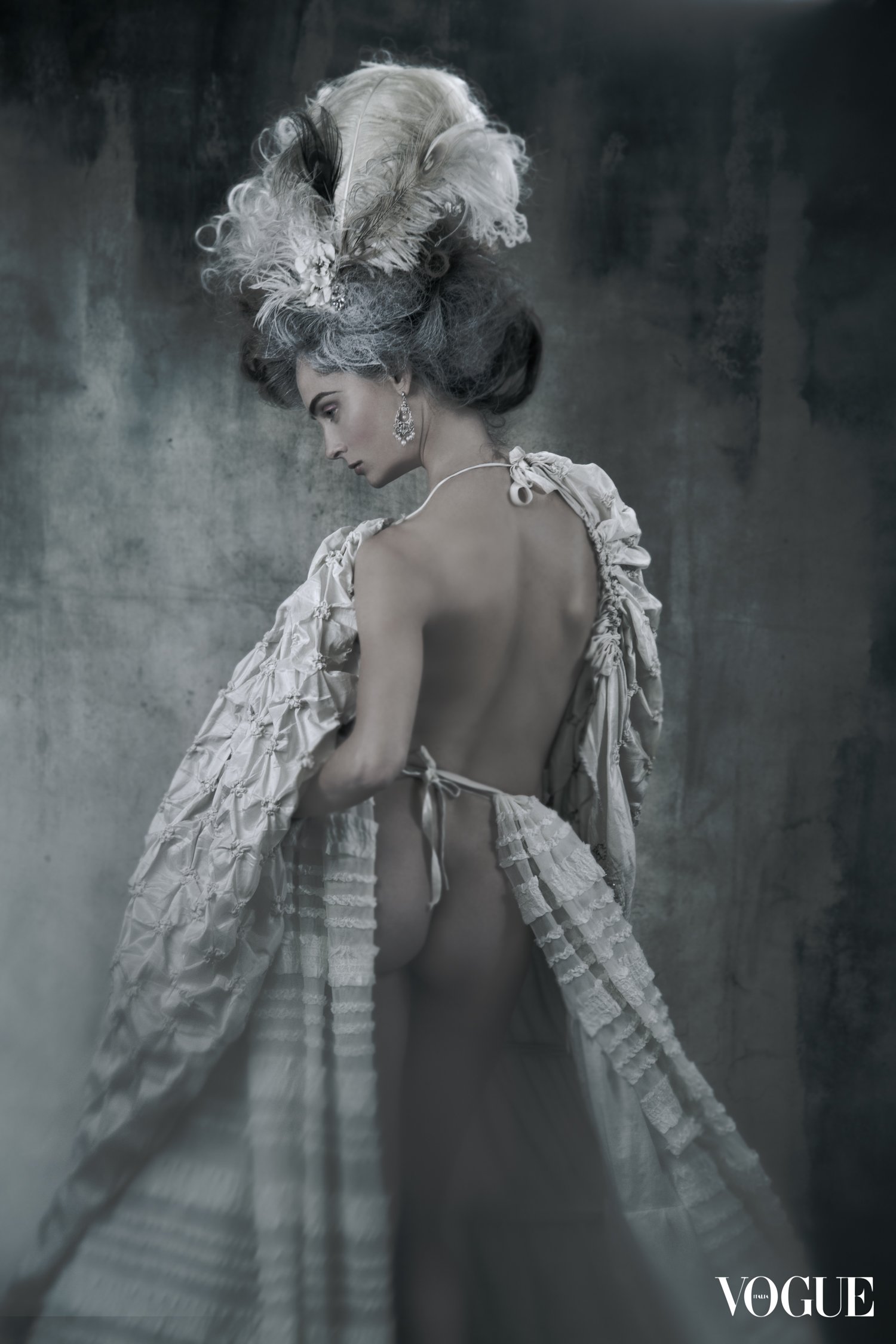 A woman with elaborate gray hair styled with feathers and jewelry, wearing a ruffled, backless gown with a tie at the waist, standing against a textured gray background, photographed for Vogue magazine.