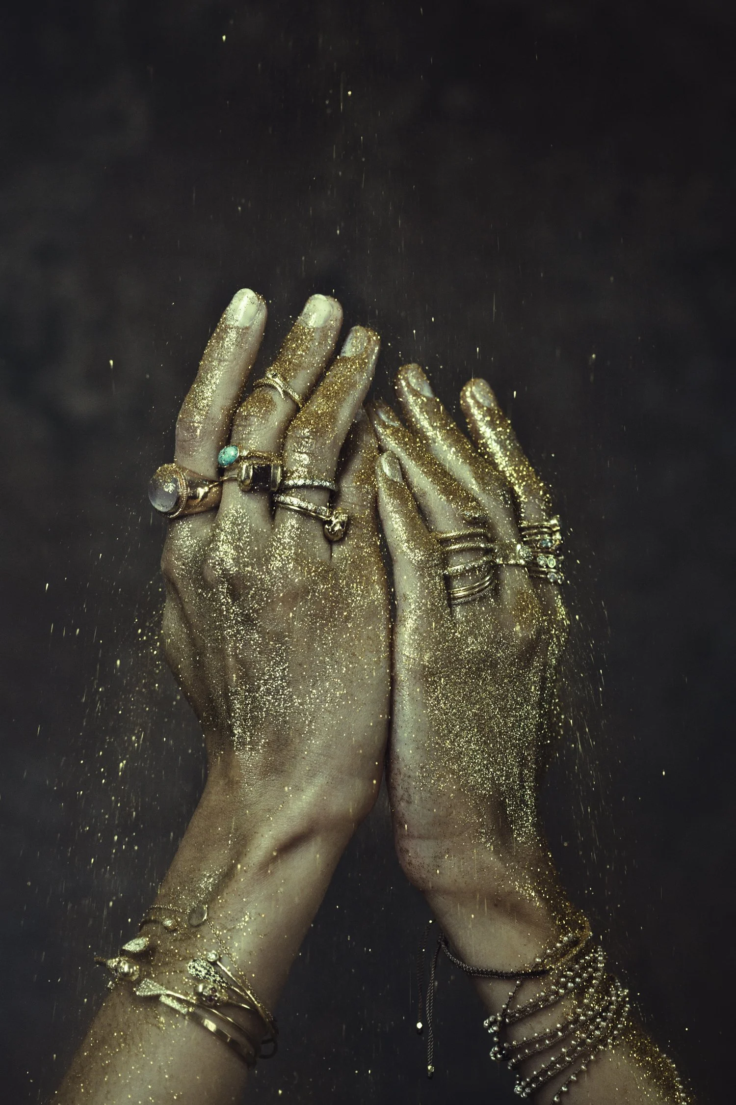 Two hands covered in gold glitter, adorned with multiple rings and bracelets, against a dark background, with glitter falling from the hands.